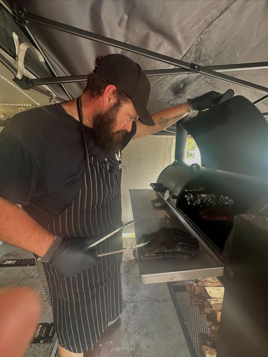 Person cooking food on a grill, wearing a black apron and gloves, in a kitchen or outdoor setting.
