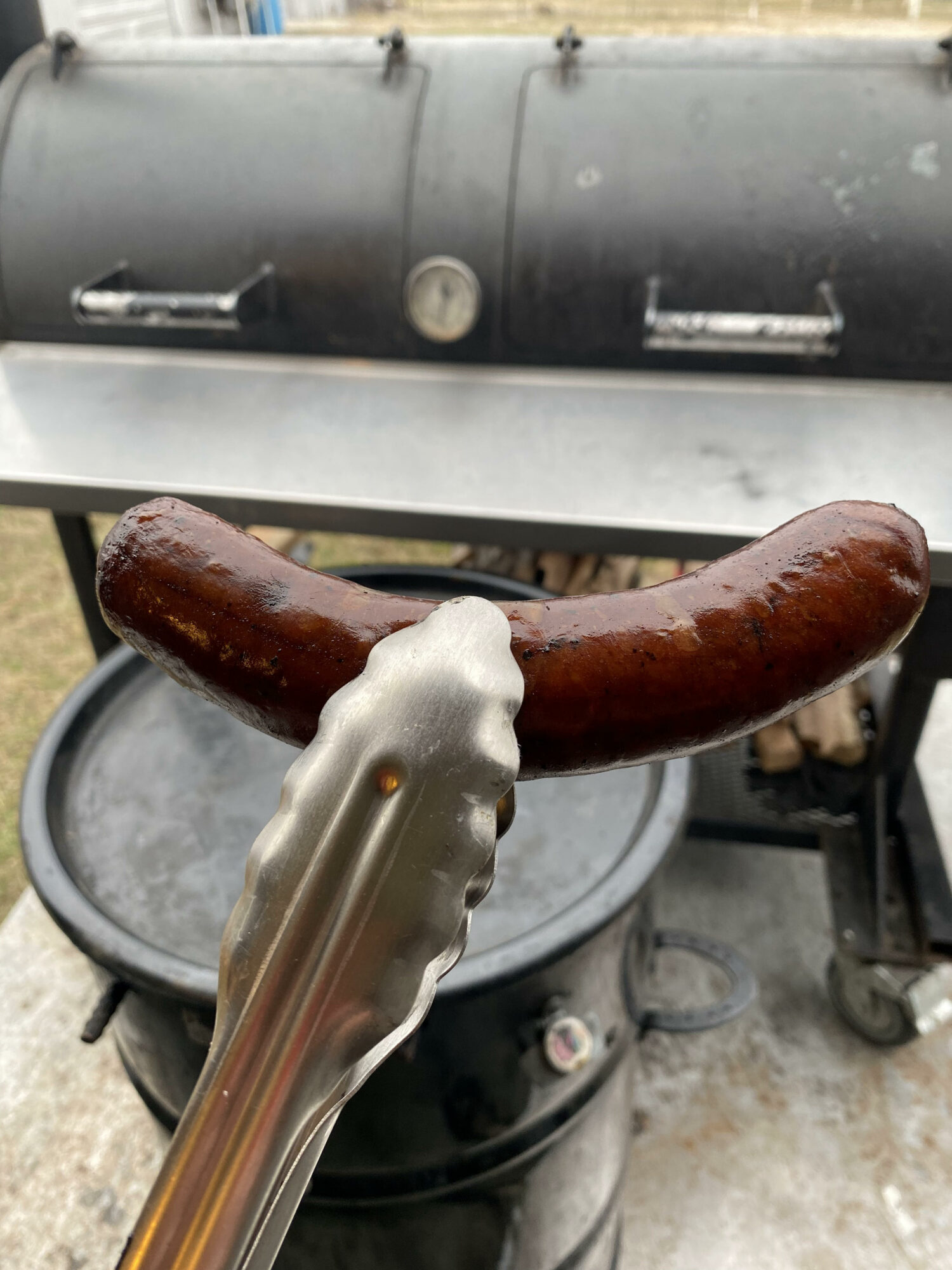 Barbecue grill with a sausage held by tongs in front of it, outdoors setting.