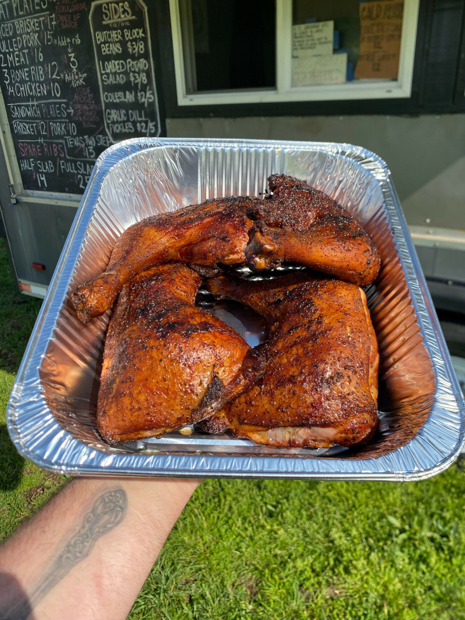 Four cooked chicken pieces in a foil tray held outdoors, with a menu board and window in the background.