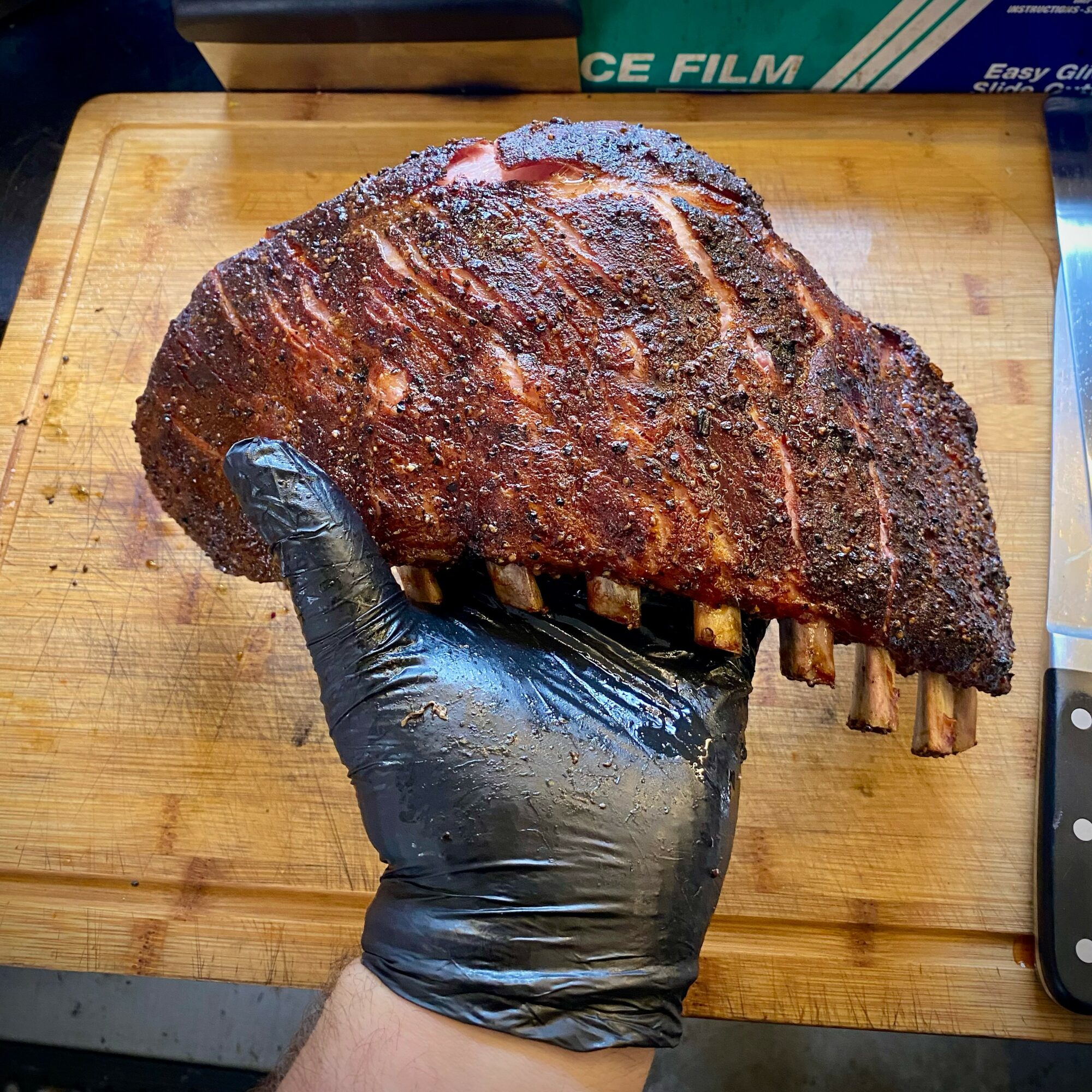 Person wearing a black glove holding a large smoked meat rib on a wooden cutting board.