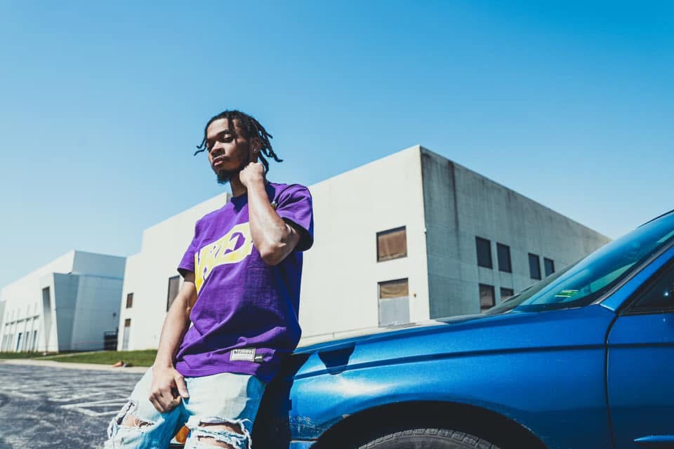 Person with dreadlocks wearing a purple and yellow sports jersey, standing next to a blue car outside a modern building.