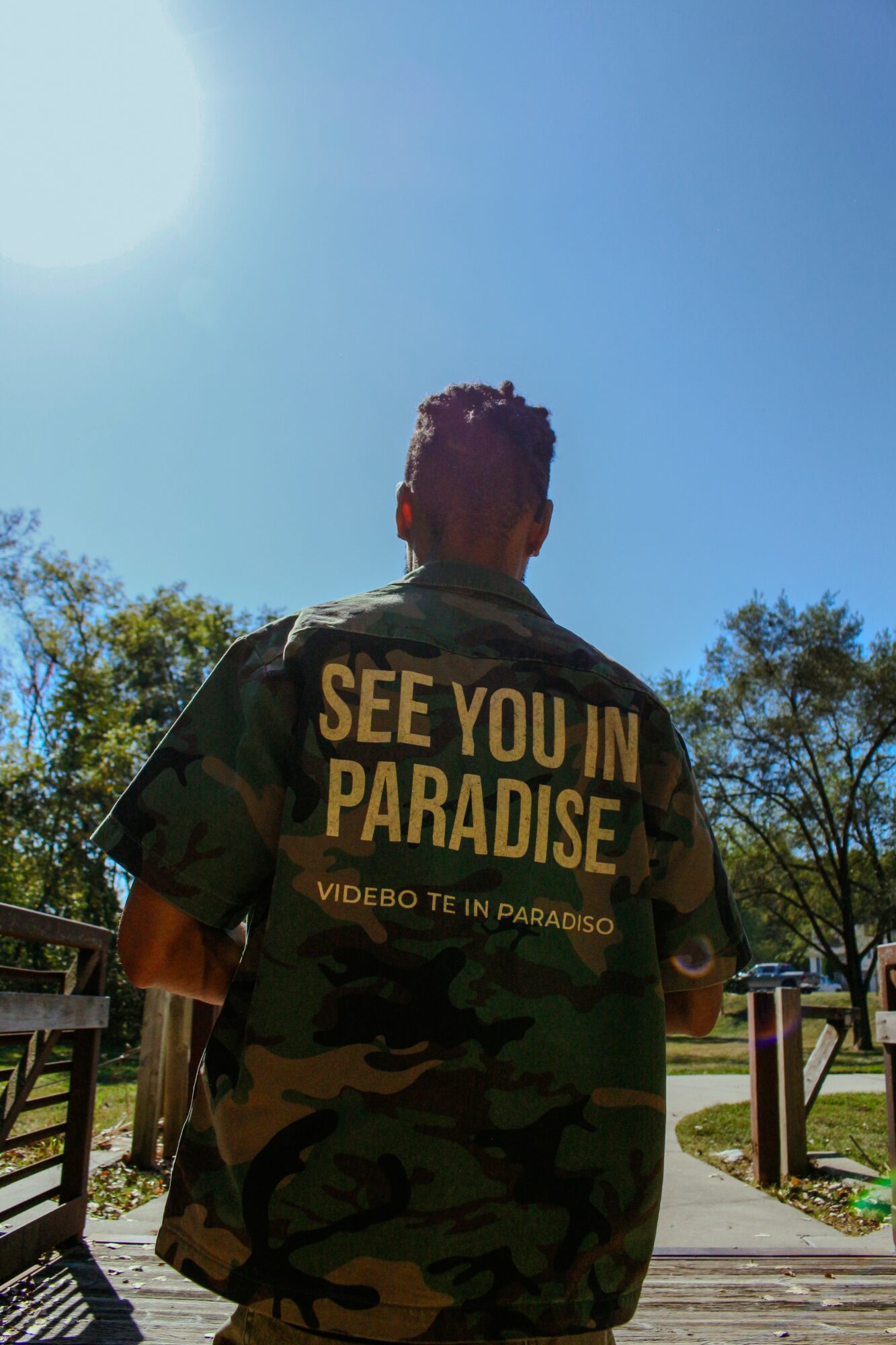 Person wearing a camouflage jacket with yellow text, standing outdoors under a bright blue sky with trees in the background.