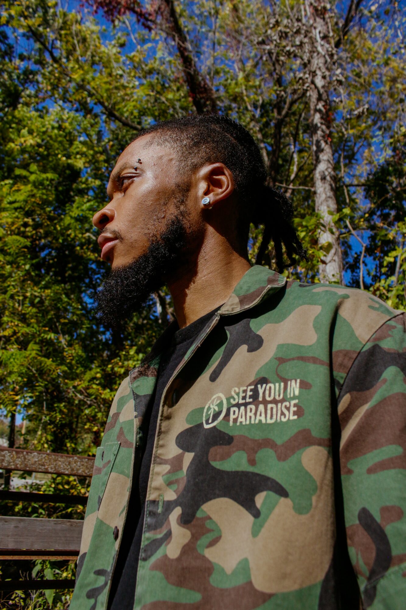 Side profile of a man with dreadlocks and beard wearing a camouflage jacket outdoors with trees and blue sky.