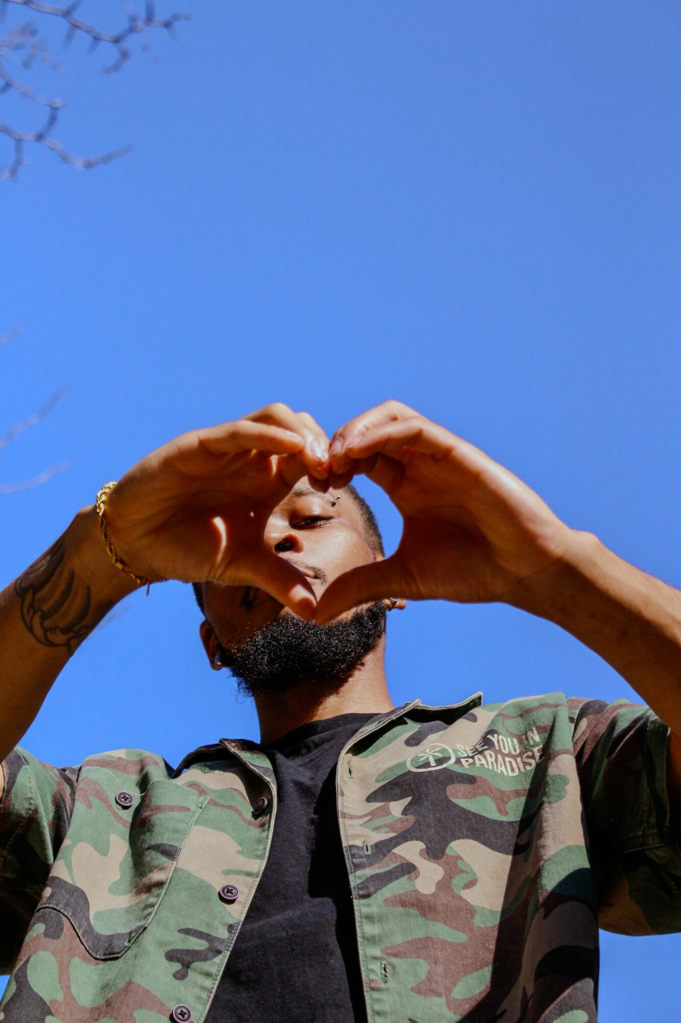 Person with a beard and tattoos making a heart shape with hands against a blue sky, wearing a camouflage jacket.