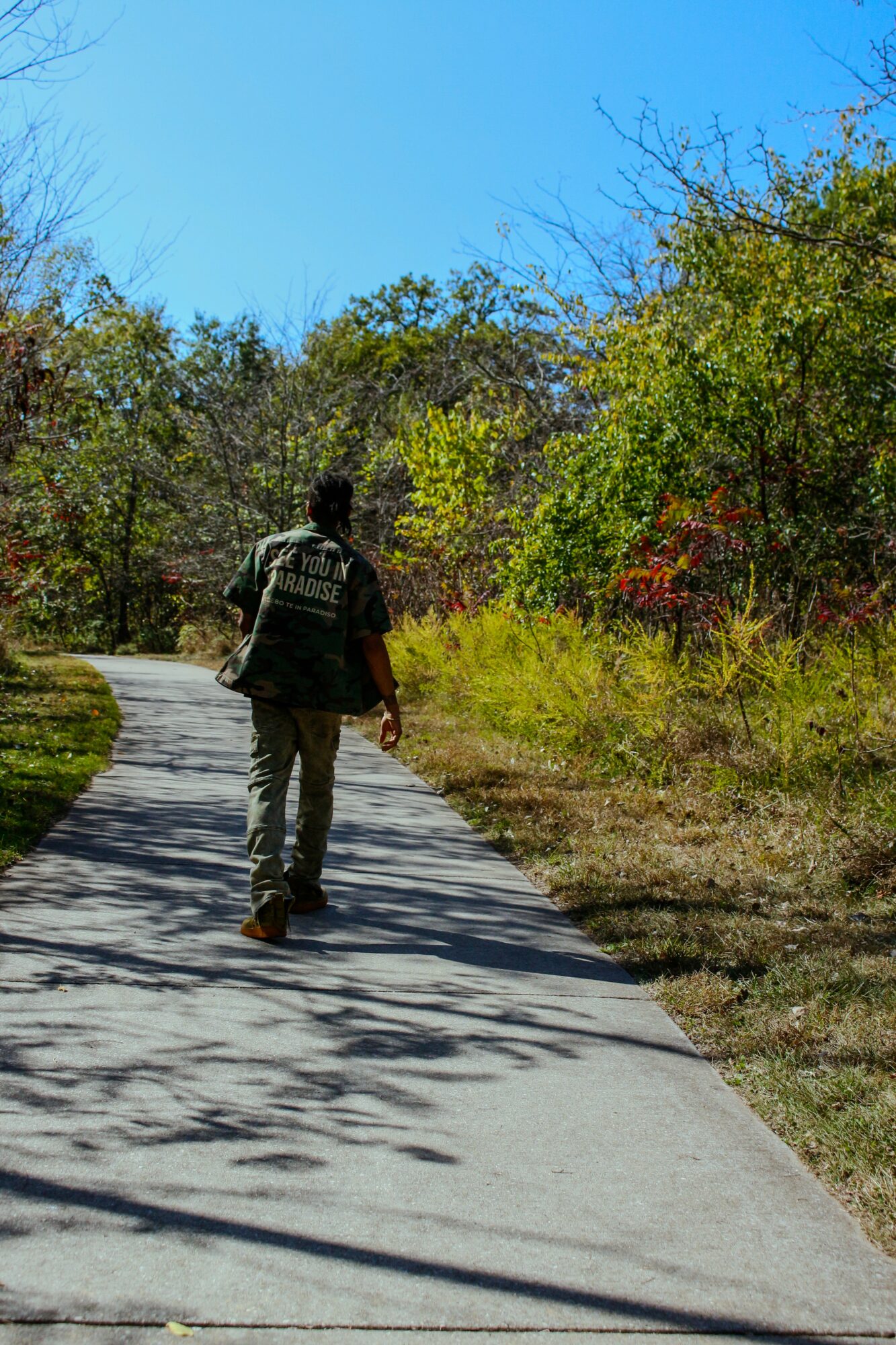 Person walking on a paved path surrounded by trees and greenery under a clear blue sky.