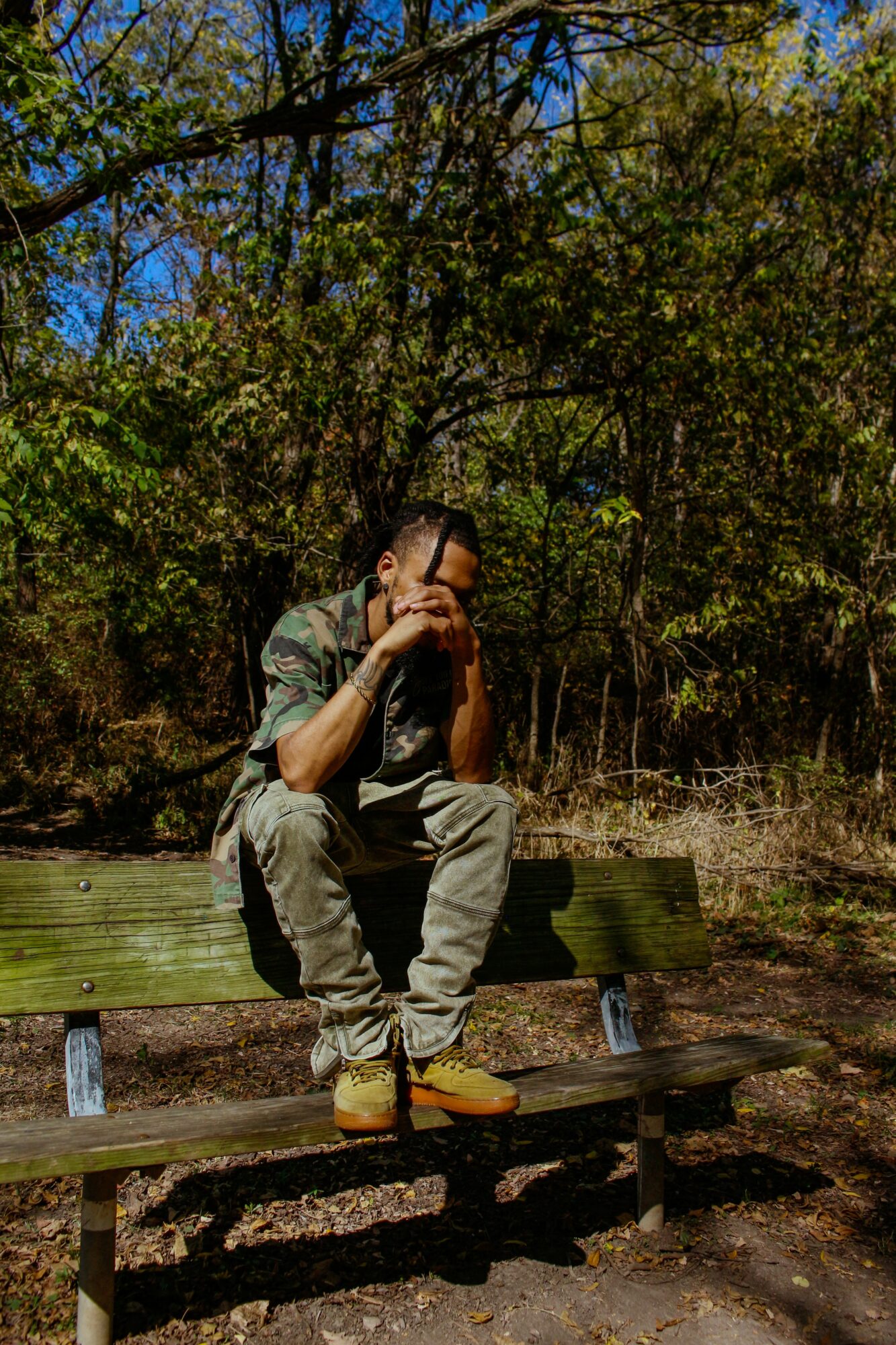 Person sitting on a park bench surrounded by trees and foliage, with hands near face, in natural outdoor setting.