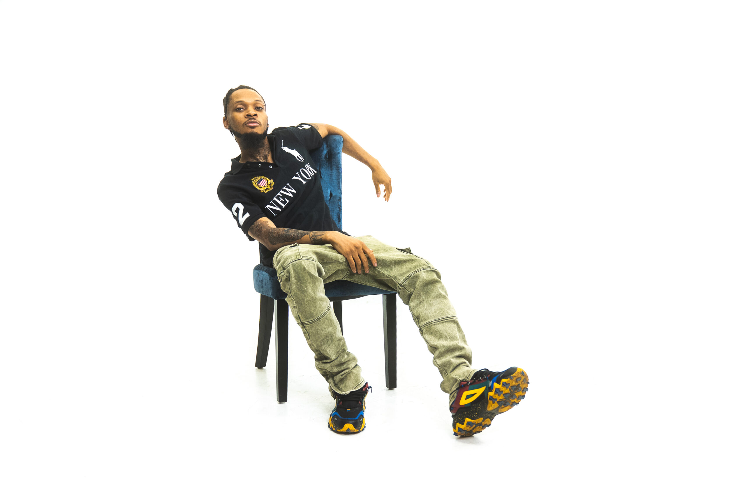 Person sitting on a chair with legs extended, wearing a black shirt, beige pants, and colorful shoes, against a white background.