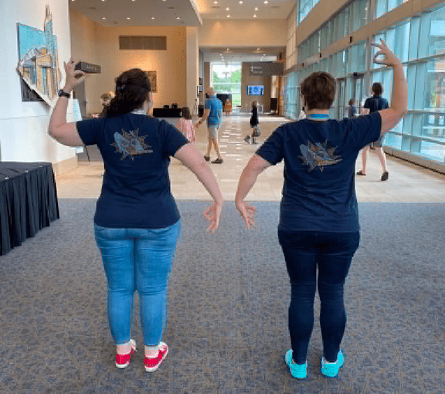 Two women in matching blue shirts and jeans walk in a spacious, well-lit indoor area with large windows and other people in the background.