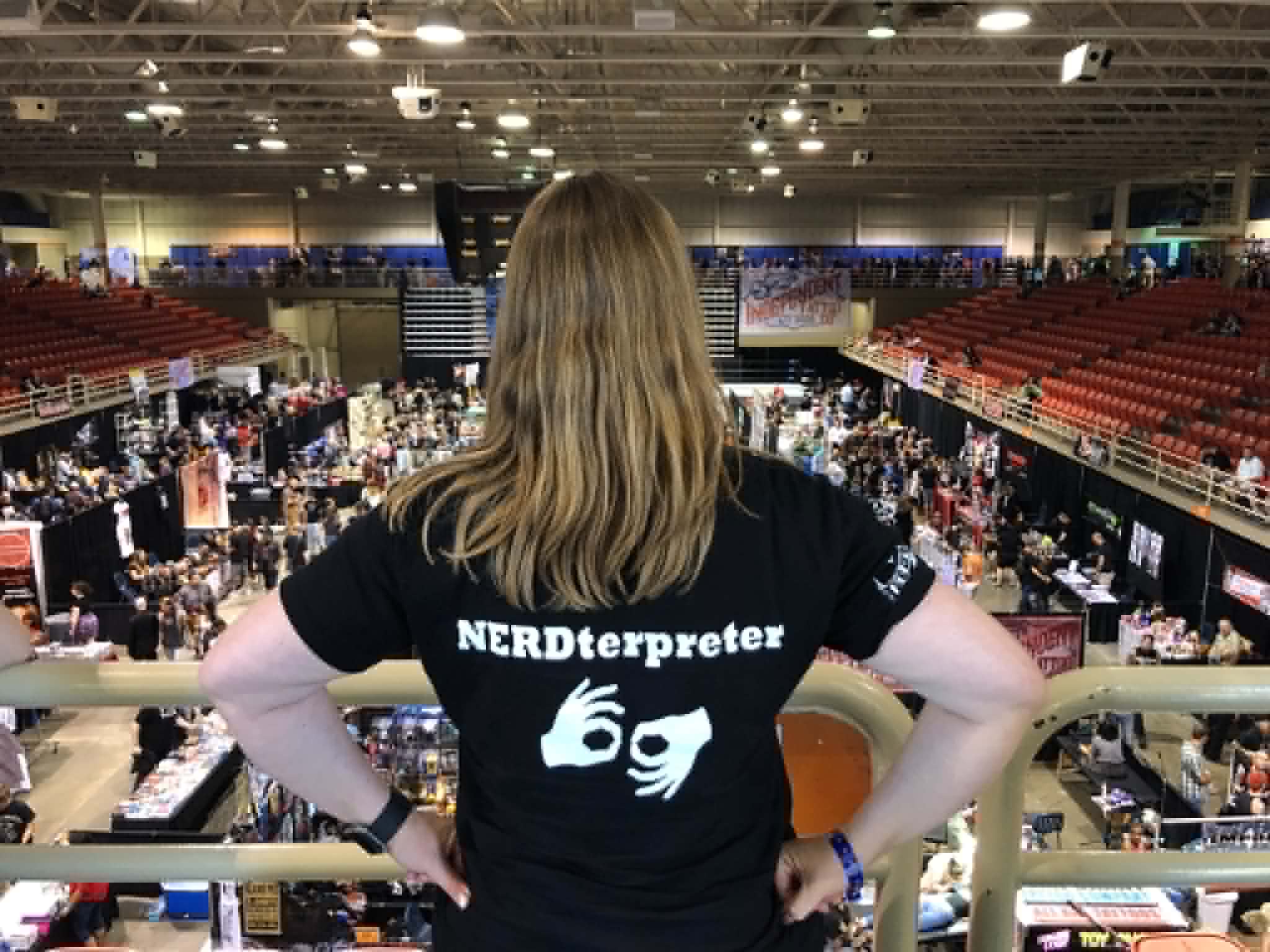 Person with long hair wearing a black T-shirt with white text, standing in an indoor event with booths and people.