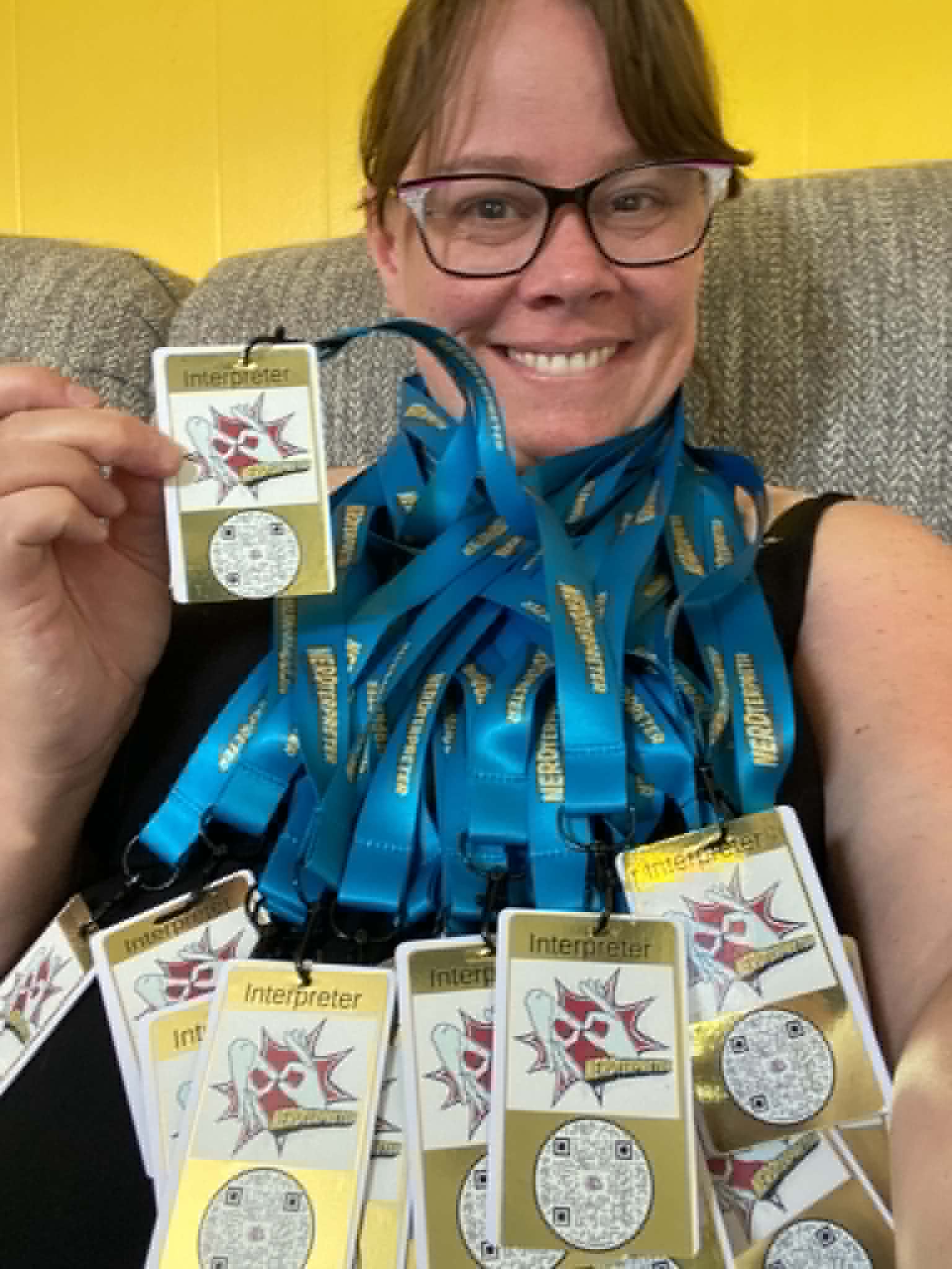 Woman with glasses smiling, holding a badge, surrounded by multiple medals, sitting on a gray couch with a yellow background.