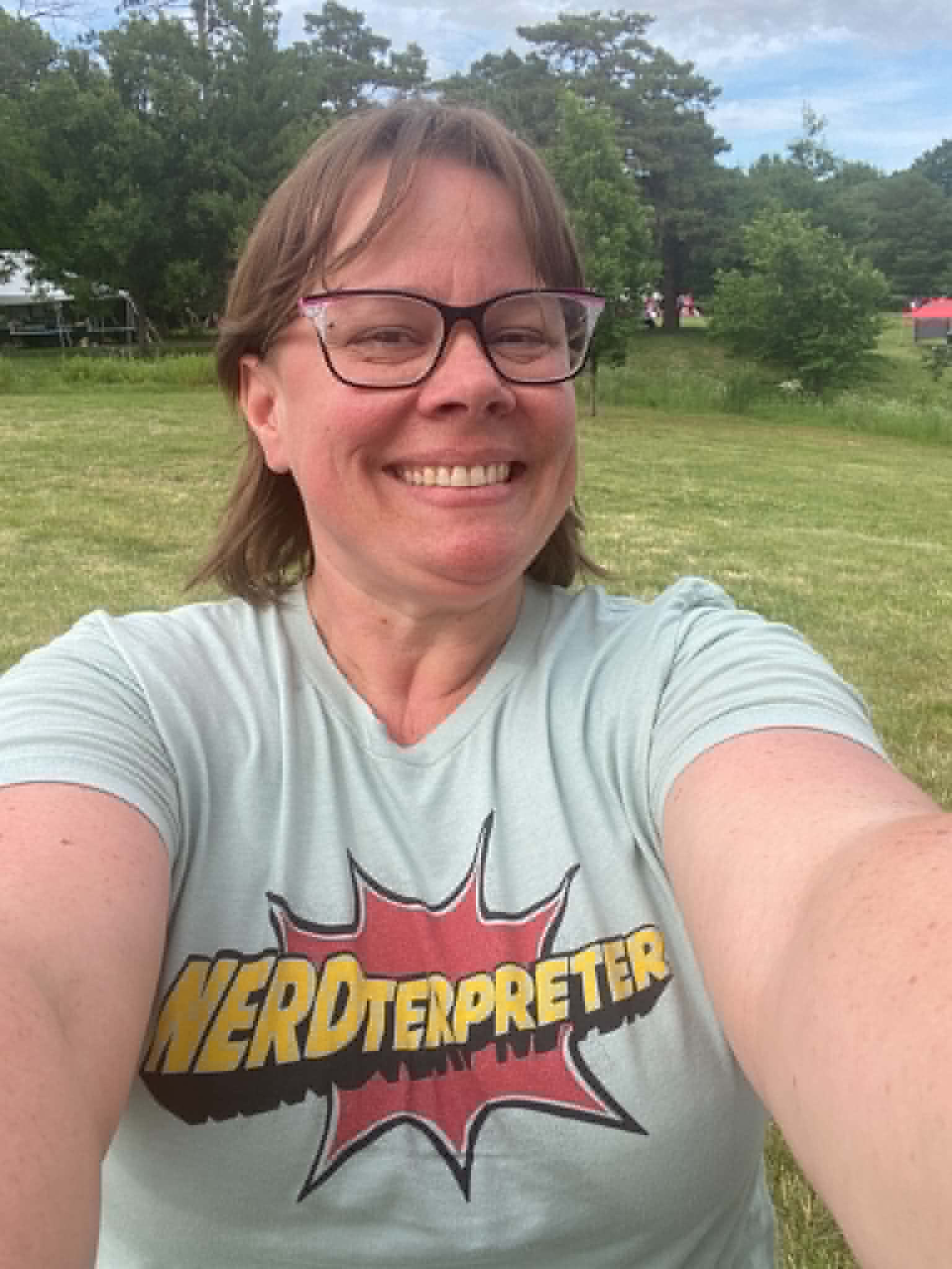 Smiling woman with glasses taking a selfie outdoors in a grassy area with trees in the background.
