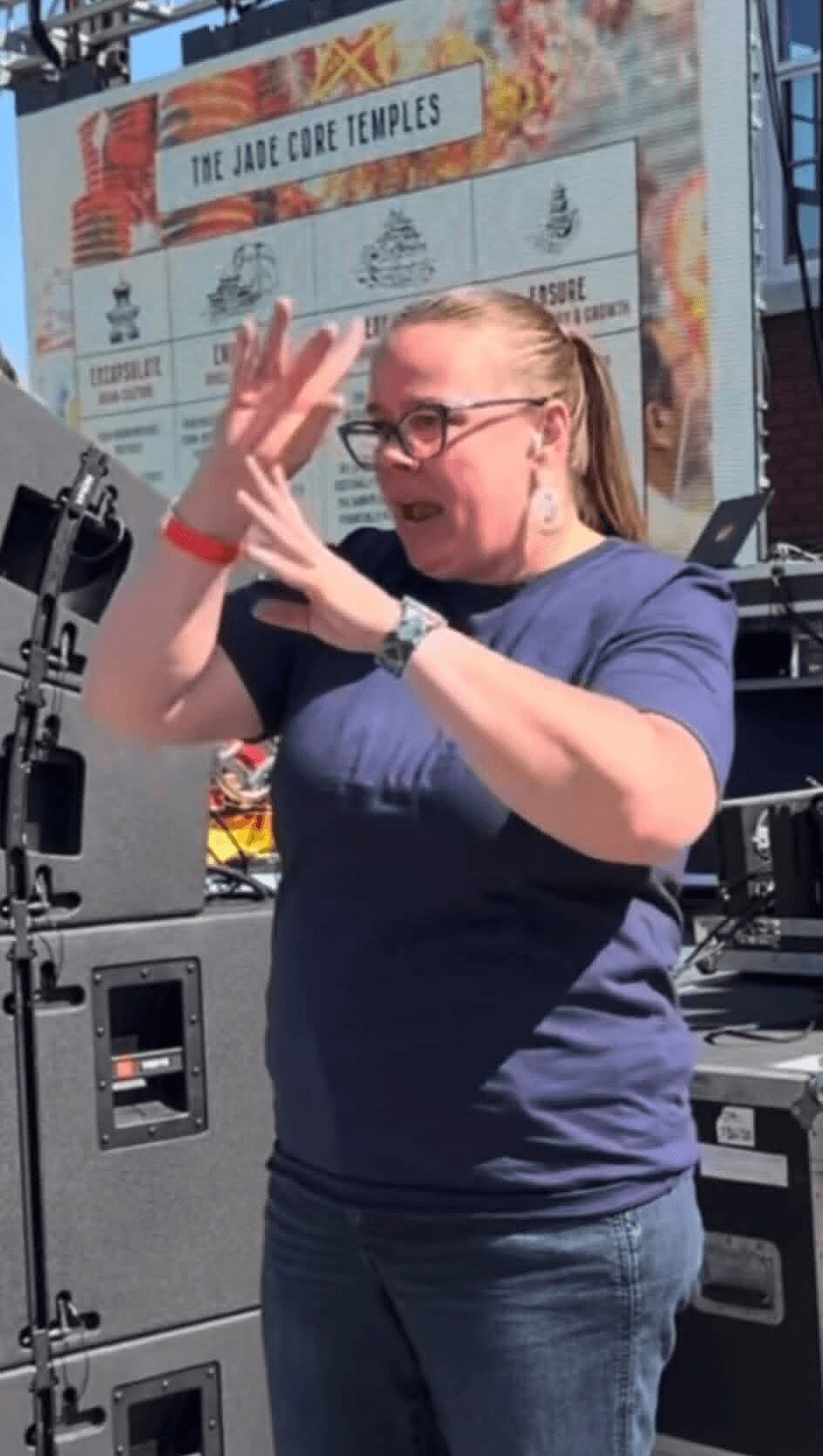 Woman with glasses and blonde hair gesturing with hands outdoors, behind sound equipment and a colorful sign.