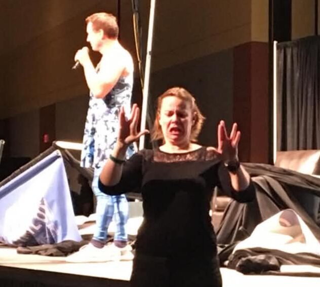 Woman speaking with hands raised, woman in background holding microphone, stage with black curtain, cluttered stage area.