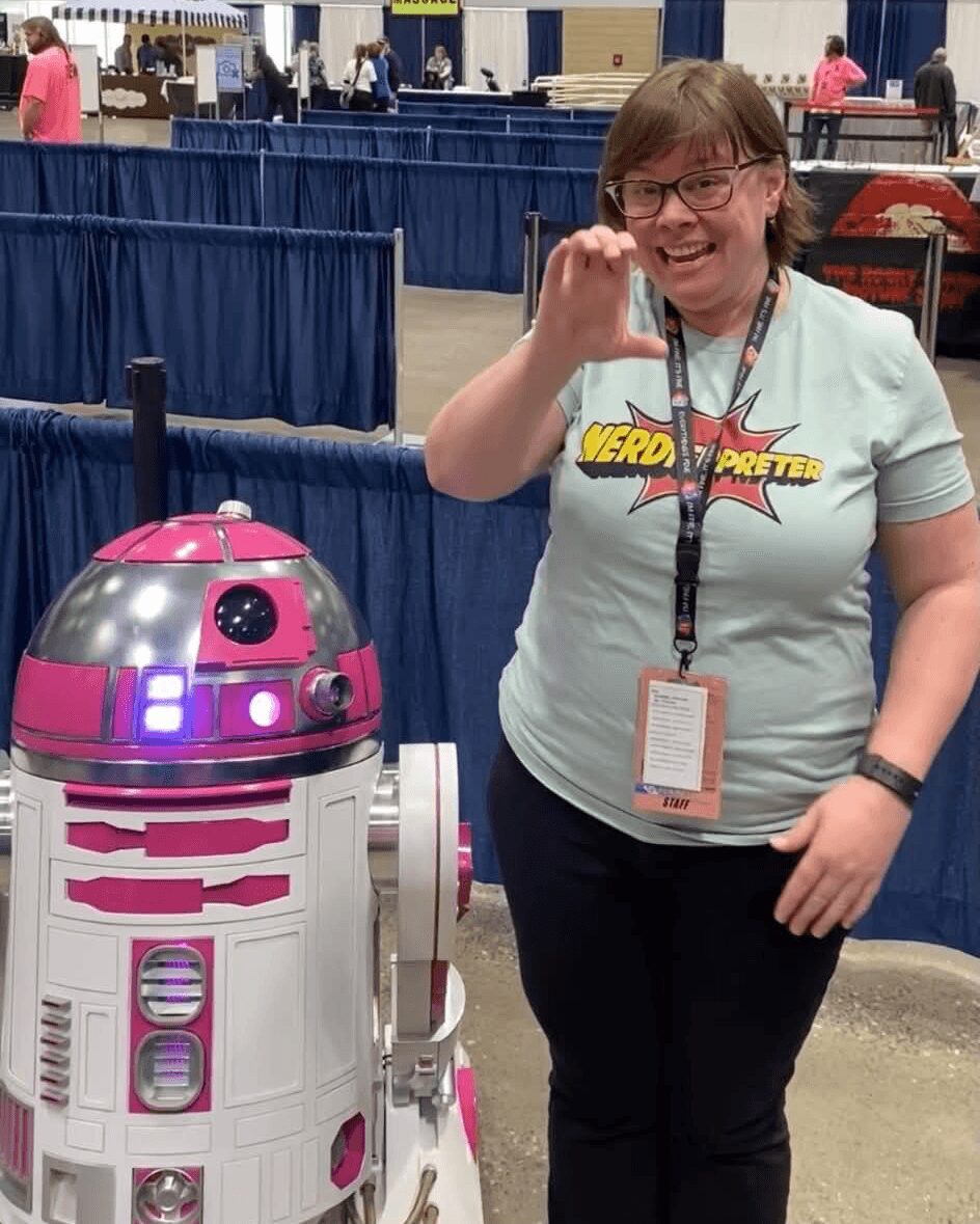 Person smiling and waving next to a pink and white robot at an indoor event.