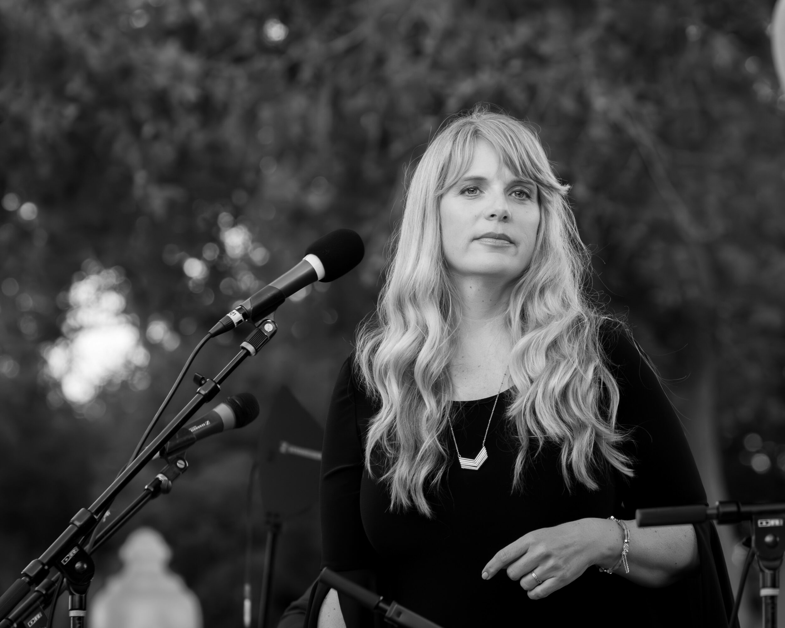 Woman with long wavy hair standing outdoors near a microphone, looking to the side, with trees in the background.