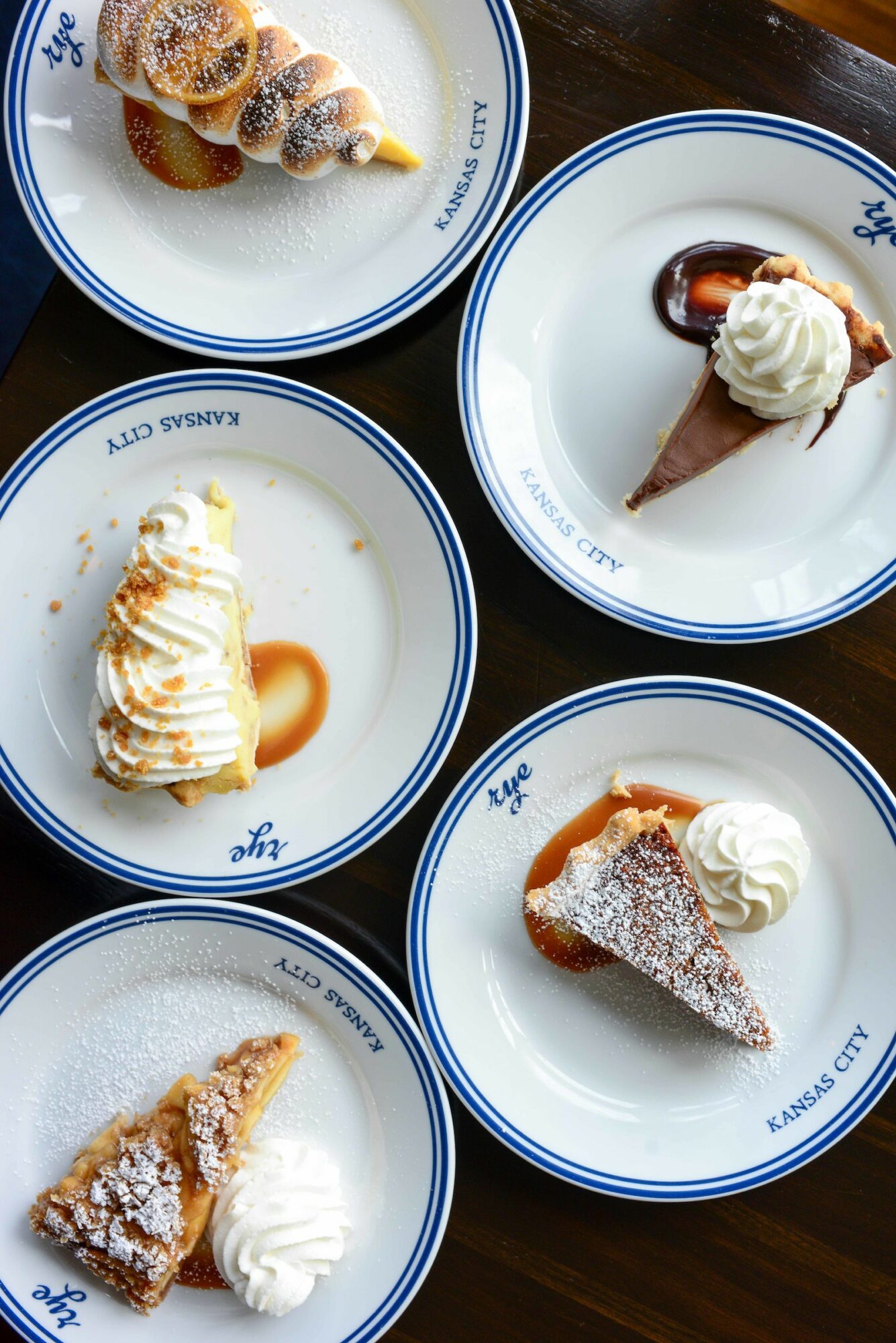 Five dessert plates with various sweets and whipped cream, arranged on a dark wooden table.
