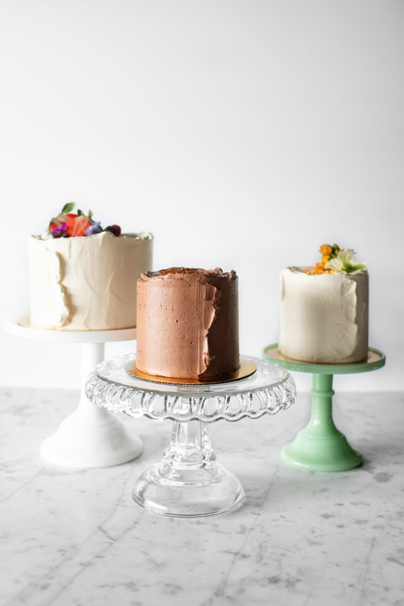 Three cakes on stands with white, chocolate, and white frosting, decorated with fruit and flowers, on a marble surface.