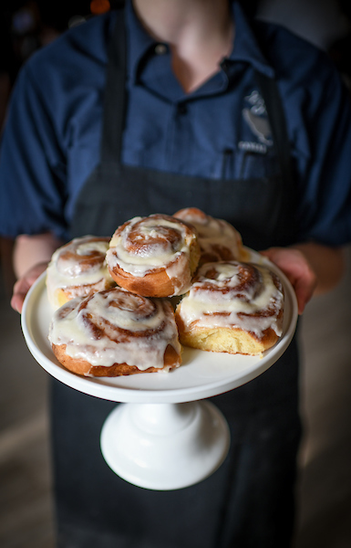Person in a blue shirt and apron holds a white cake stand with cinnamon rolls topped with icing.