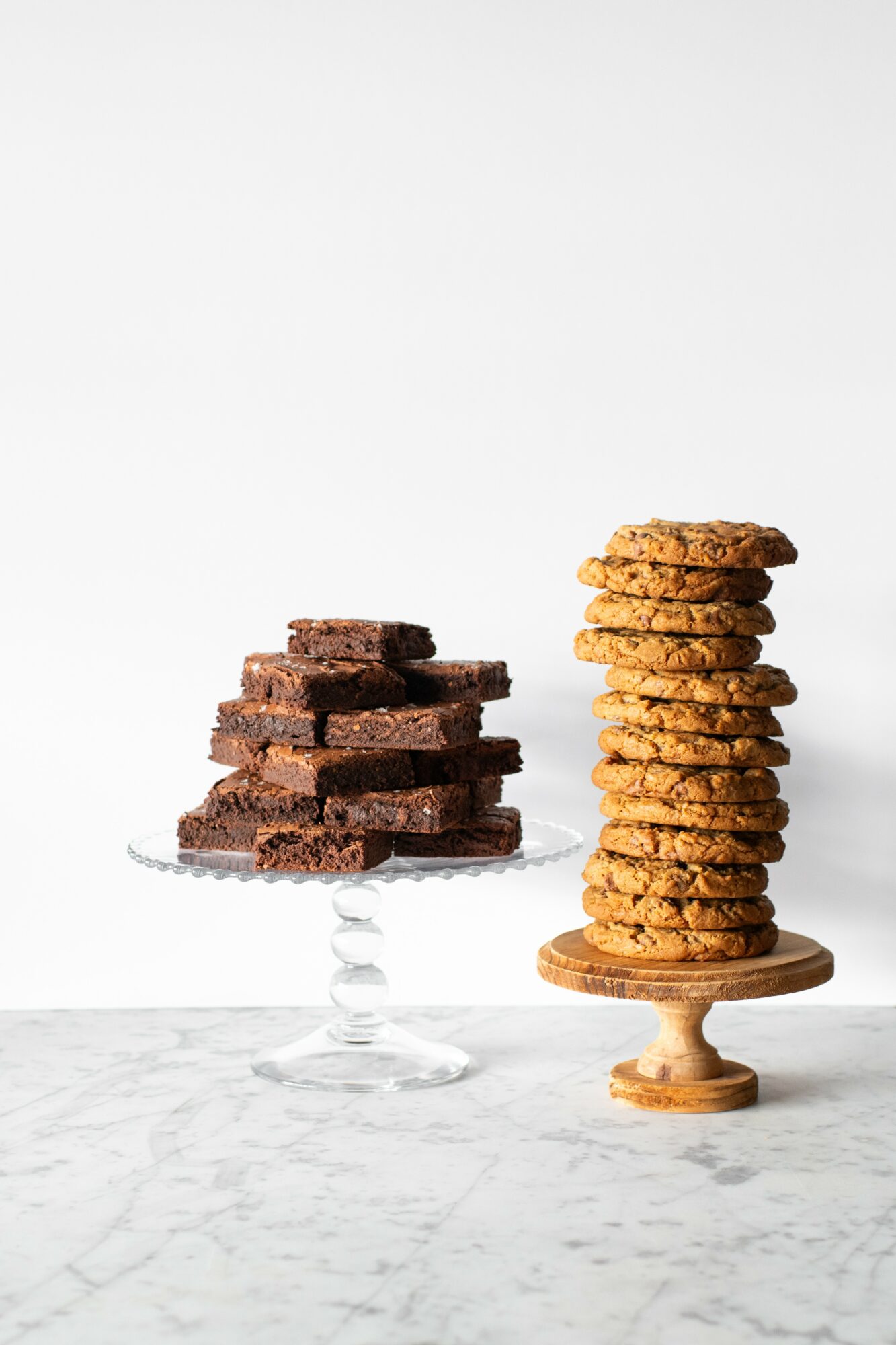 Stack of chocolate brownies on a glass cake stand and a stack of cookies on a wooden stand, both on a white surface.