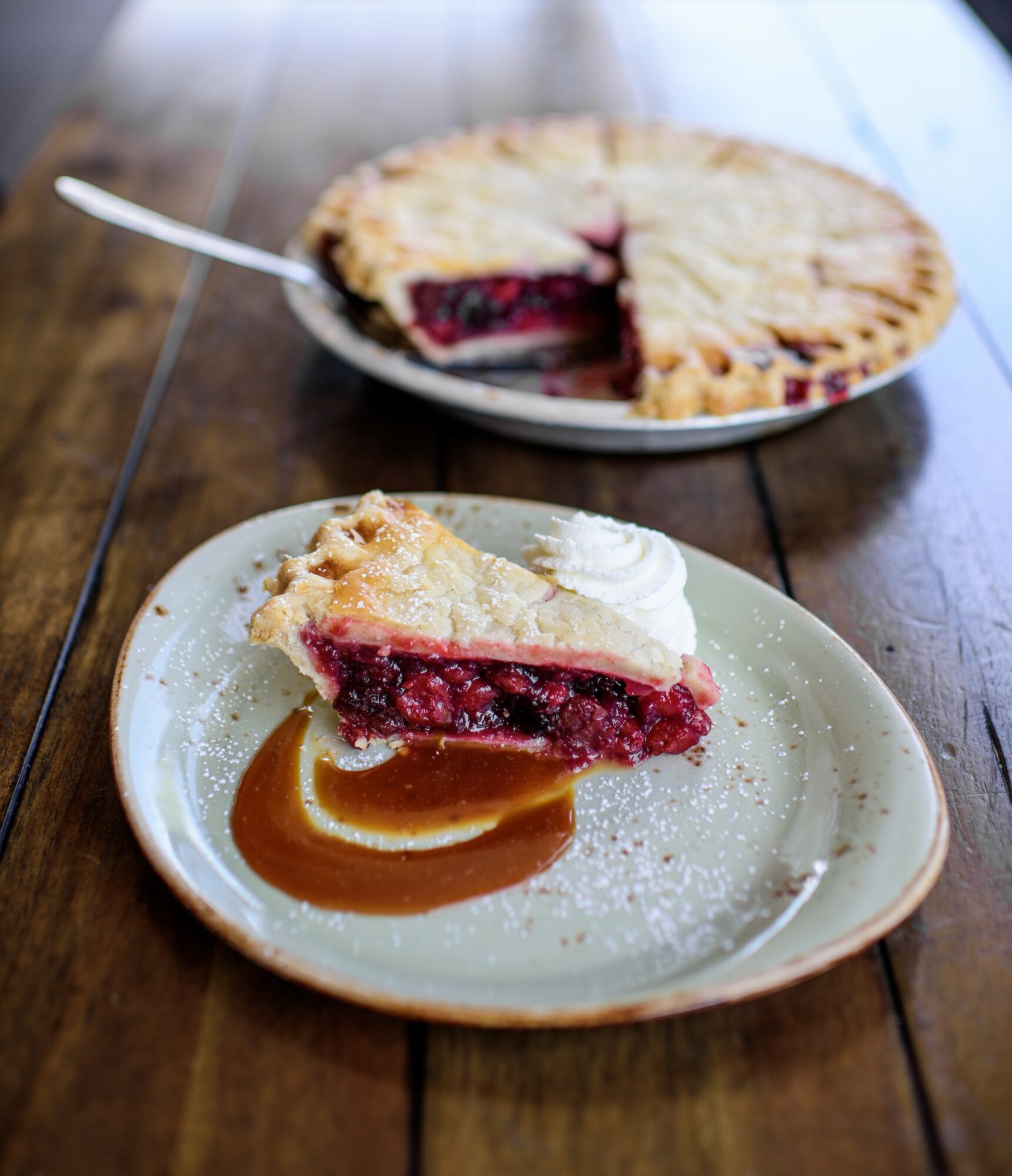 Slice of cherry pie on a plate with caramel sauce, with the remaining pie in the background on a wooden table.