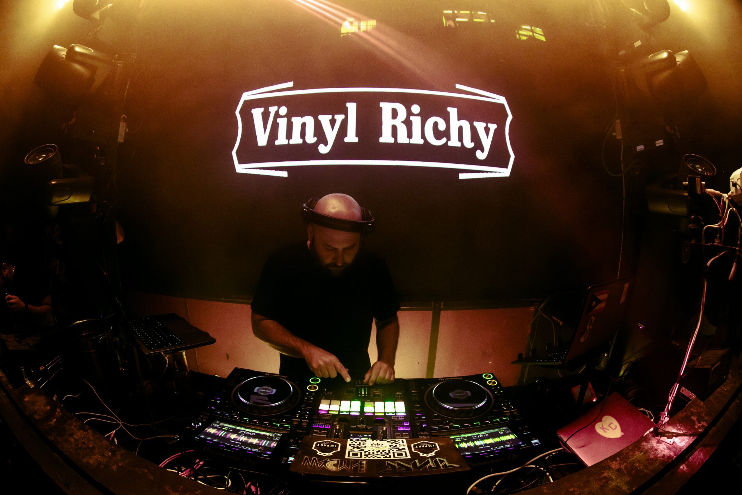DJ wearing a hat operating turntables in front of a screen displaying 'Vinyl Richy' at a music event.