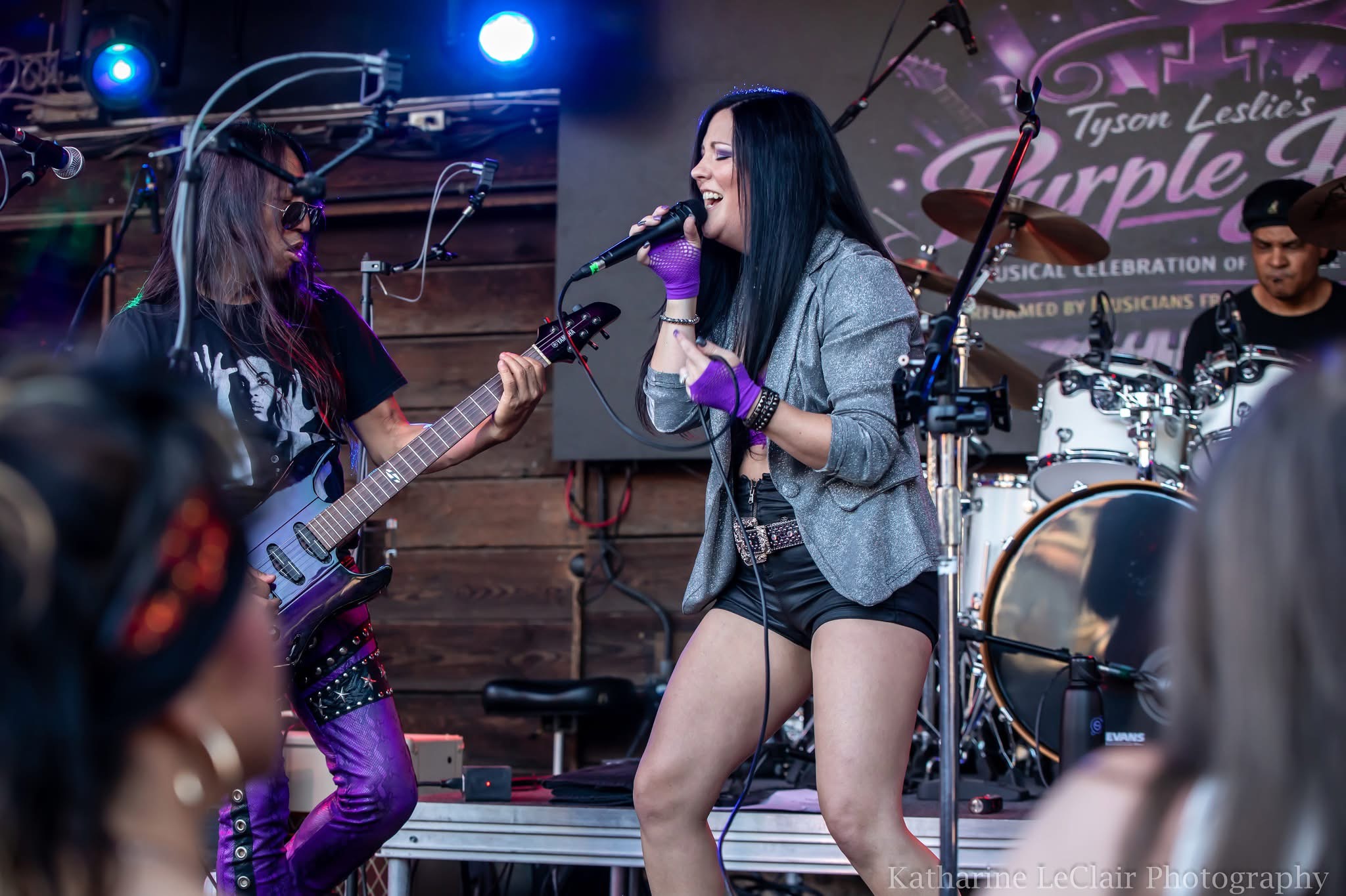 Female singer with long dark hair performs with a band on stage, playing guitar and drums in the background.