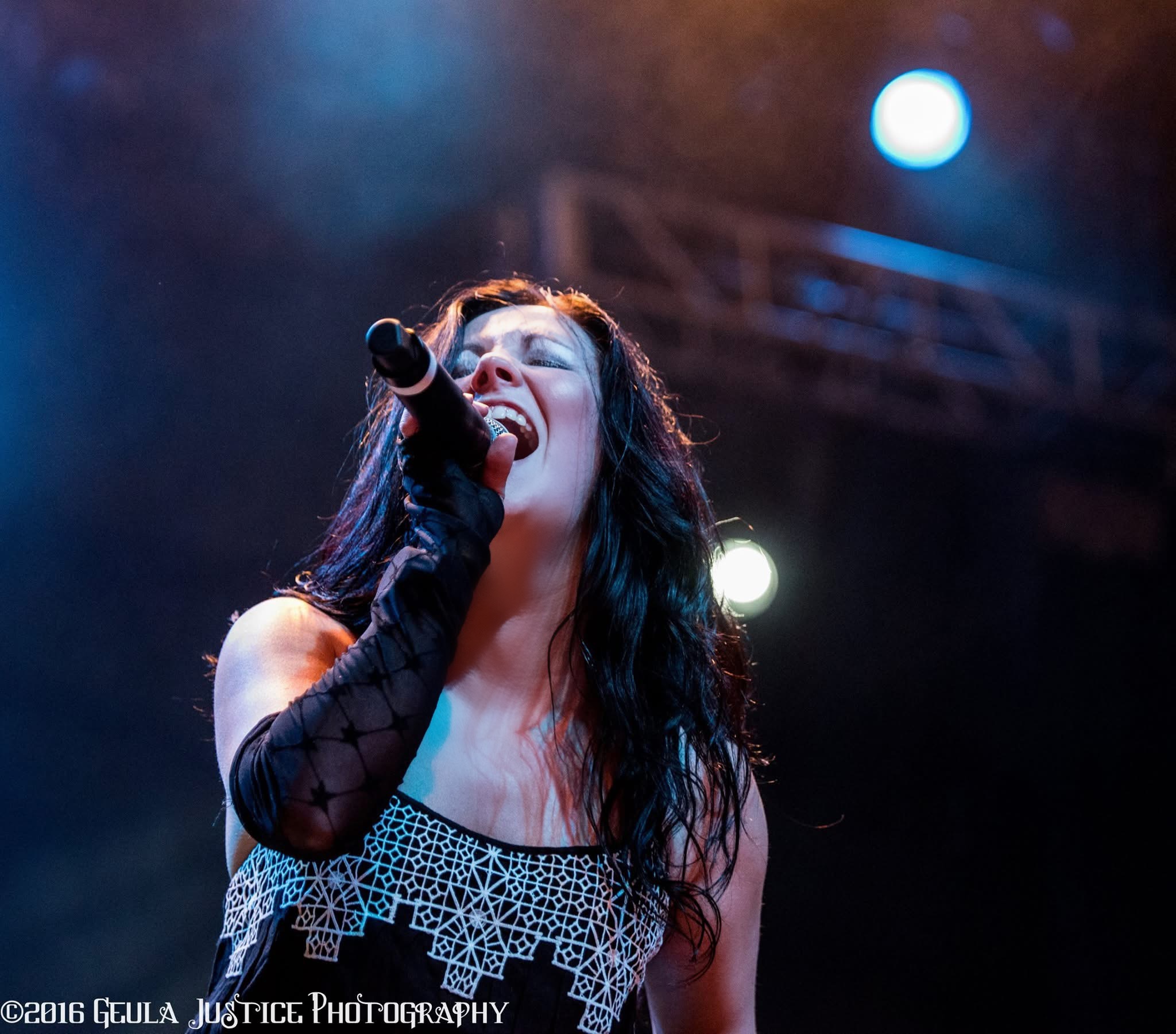 Female singer with long dark hair performs on stage, holding a microphone, with stage lights in background.