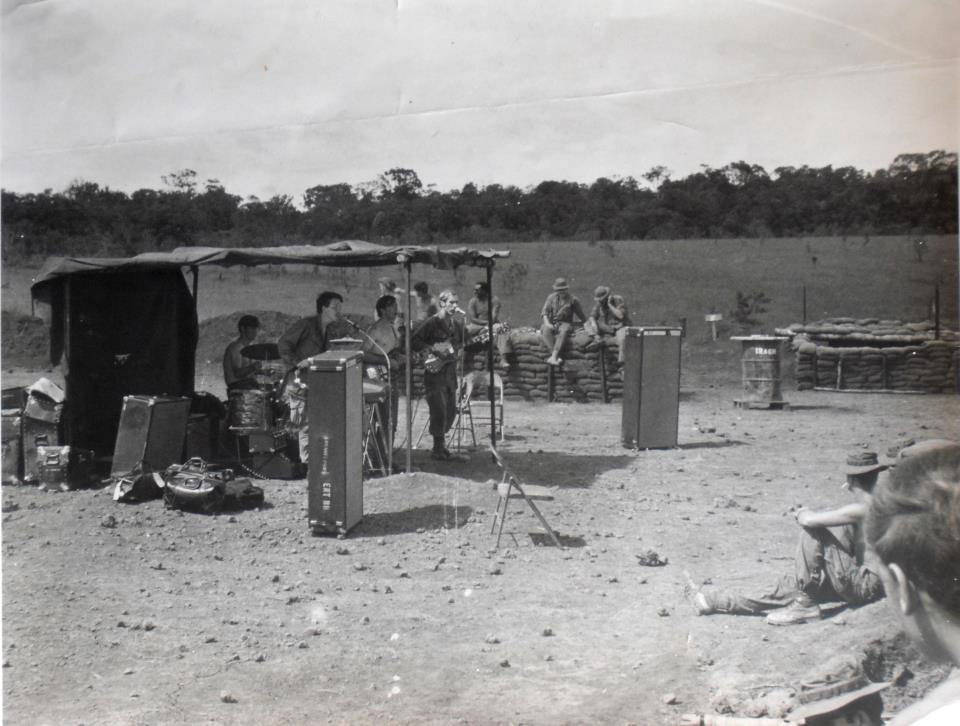 Group of people outdoors near a makeshift shelter with equipment, some sitting and others standing, in a rural setting.