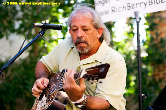 Man playing guitar outdoors with microphone and banner in background, trees and sunlight visible