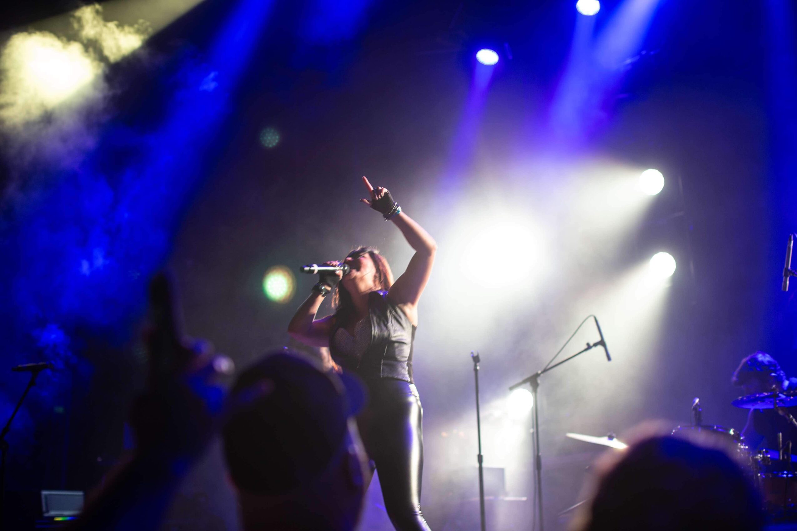 Performer singing on stage with bright lights and audience in foreground, pointing upward with one hand.