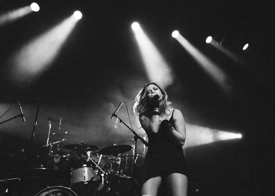 Female singer performing on stage with drums and bright stage lights behind her, in black and white.