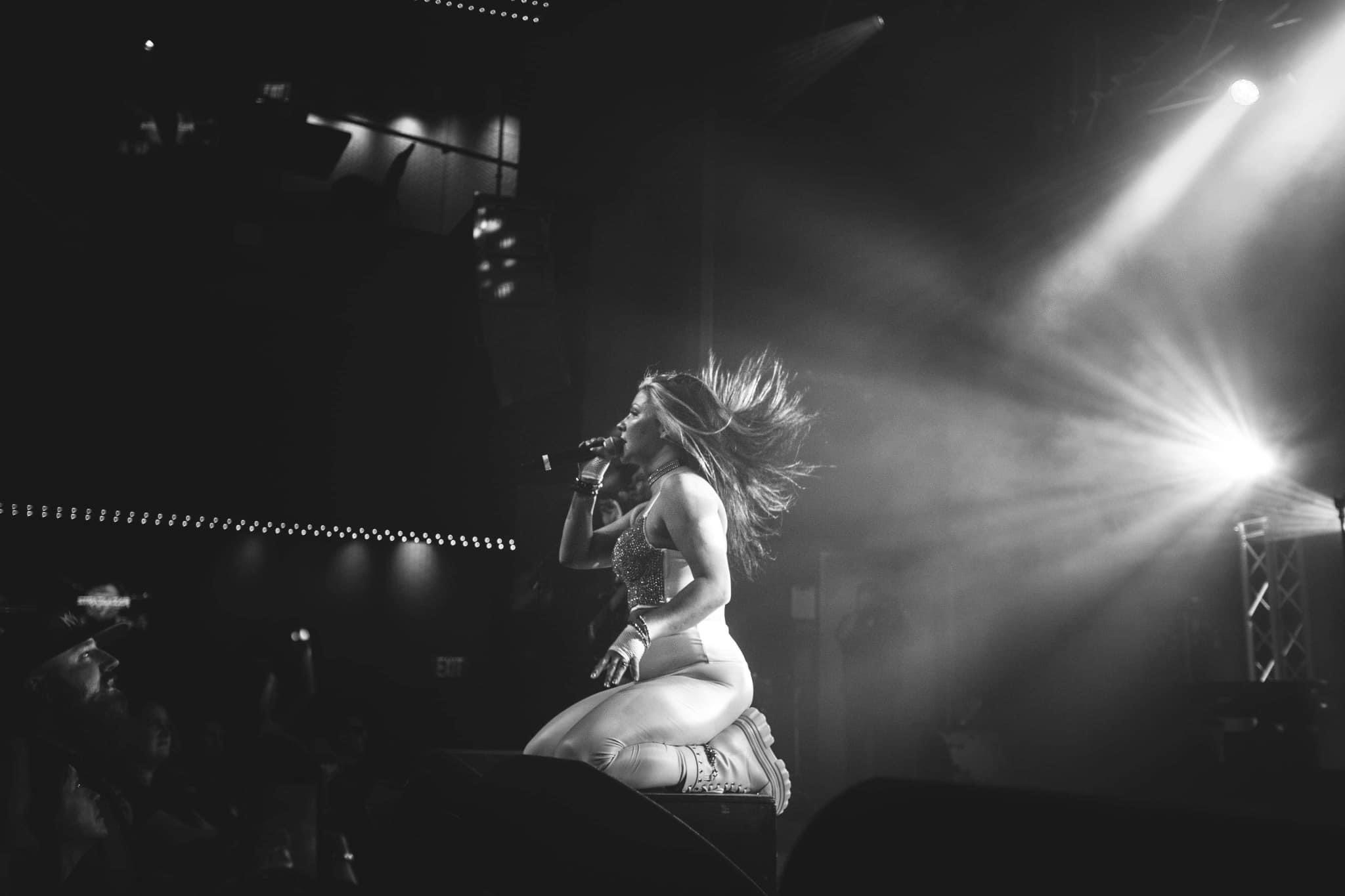 A woman with long hair is singing into a microphone on stage with bright lights shining behind her.