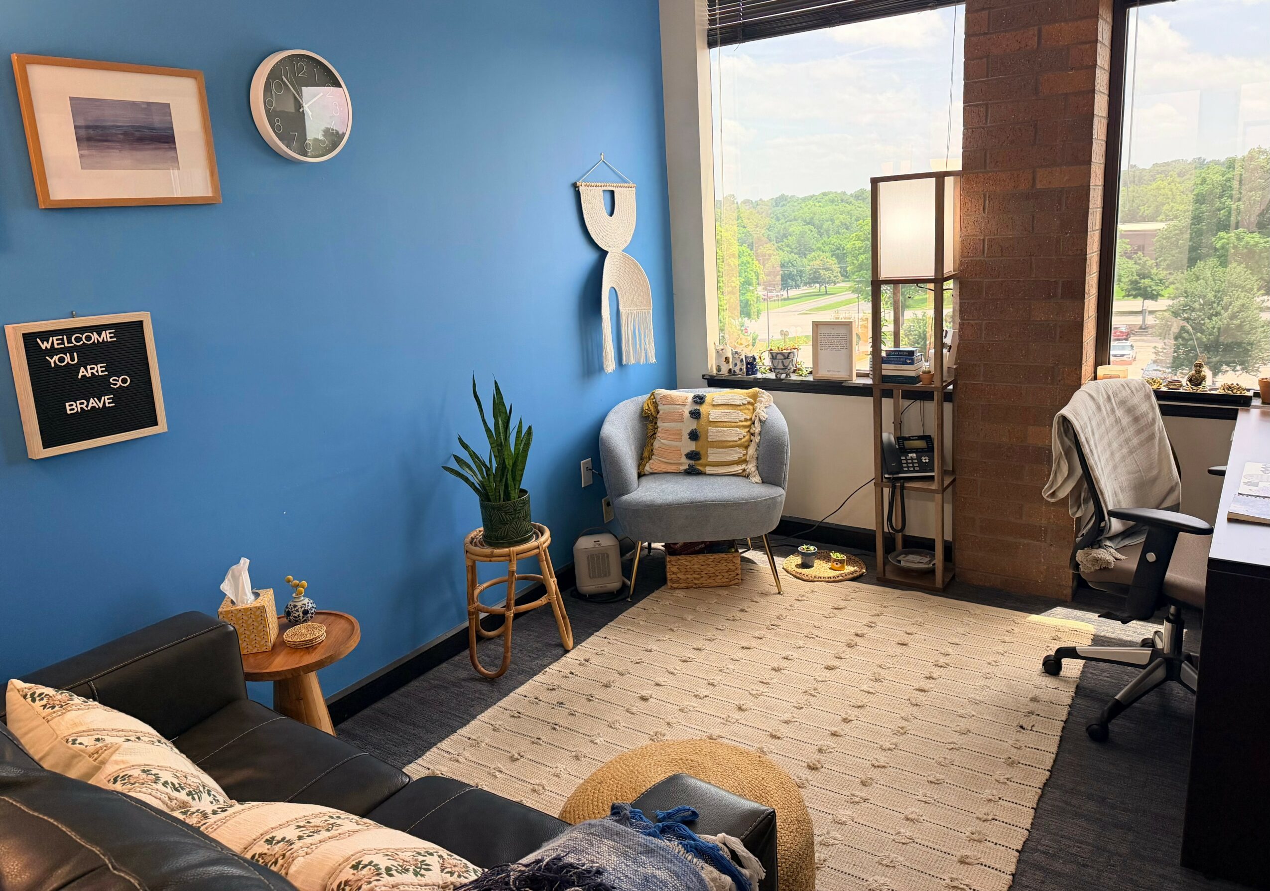 Office with blue wall, window view, sofa, armchair, plant, and desk, decorated with wall art and a rug.