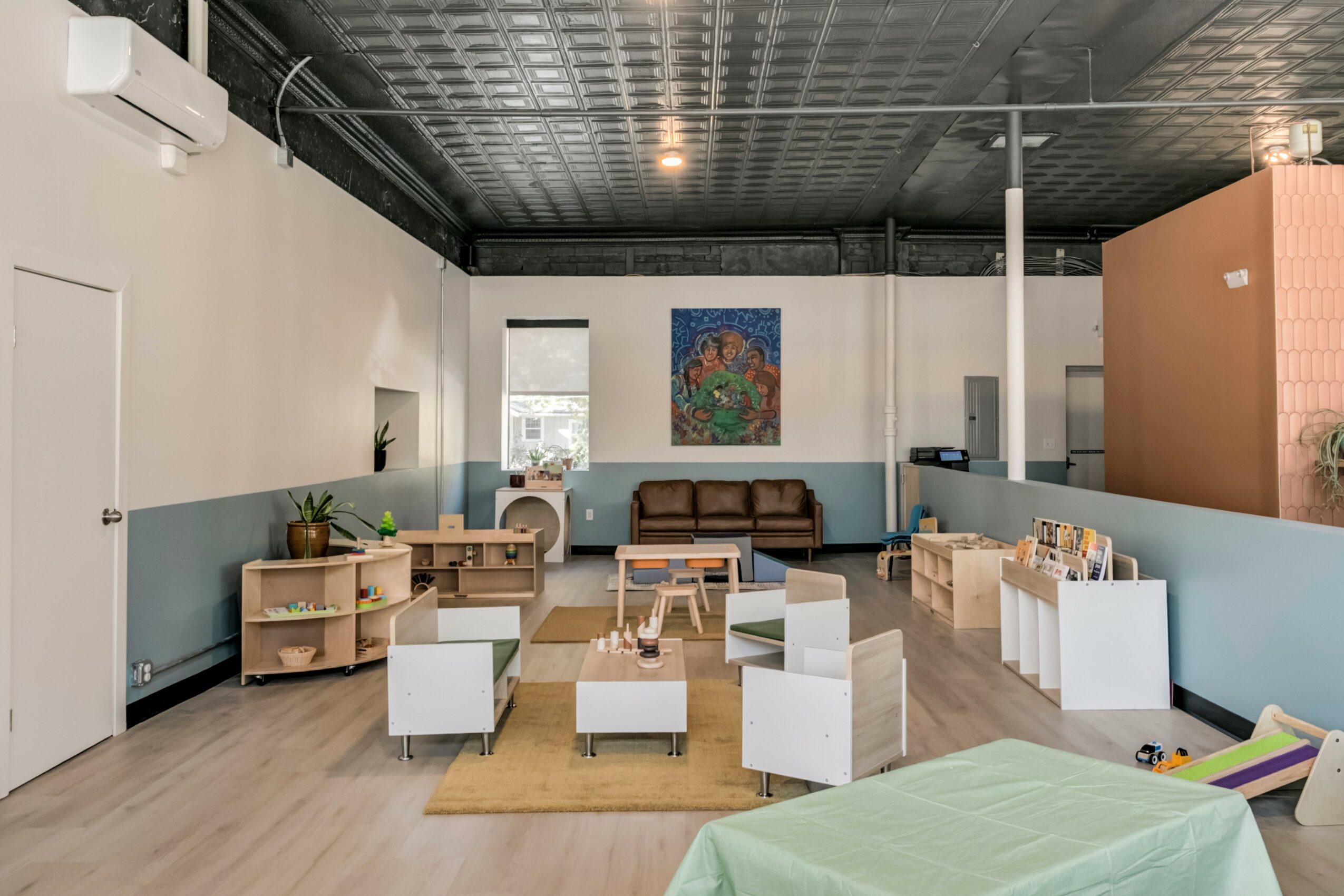 Children's playroom with chairs, tables, and a brown sofa, decorated with a colorful painting on the wall.