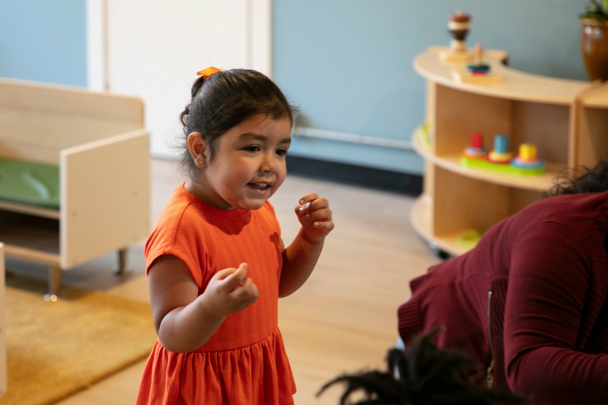 Young girl in orange dress smiling and gesturing indoors, with a wooden shelf and play area in background.