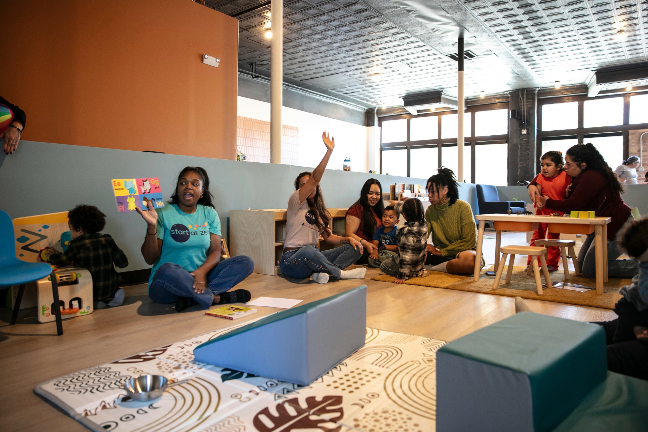 Children and adults sitting on the floor in a room with large windows, some raising hands, engaging in activities.