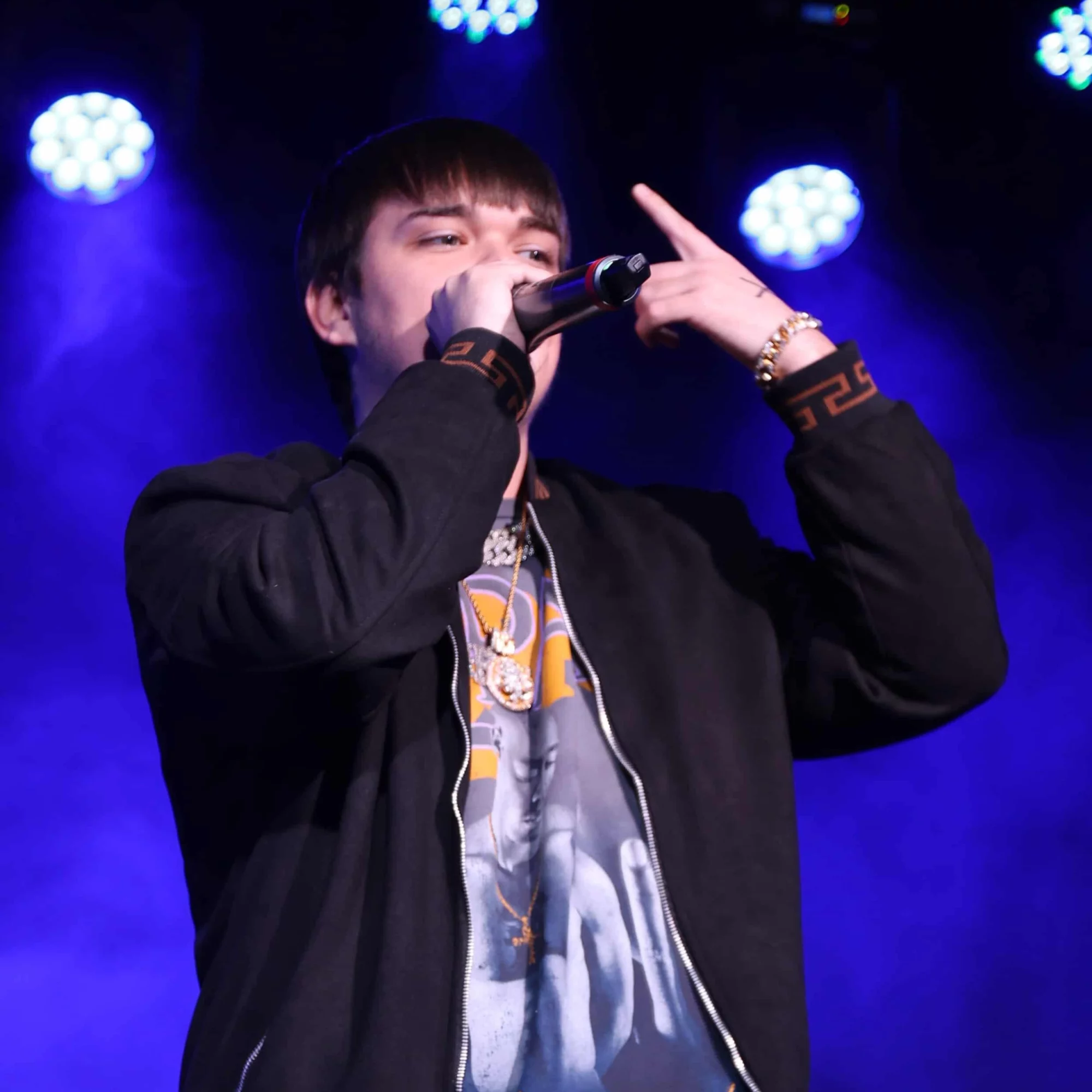 Young man performing on stage with blue lights, holding a microphone, wearing a black jacket and jewelry.