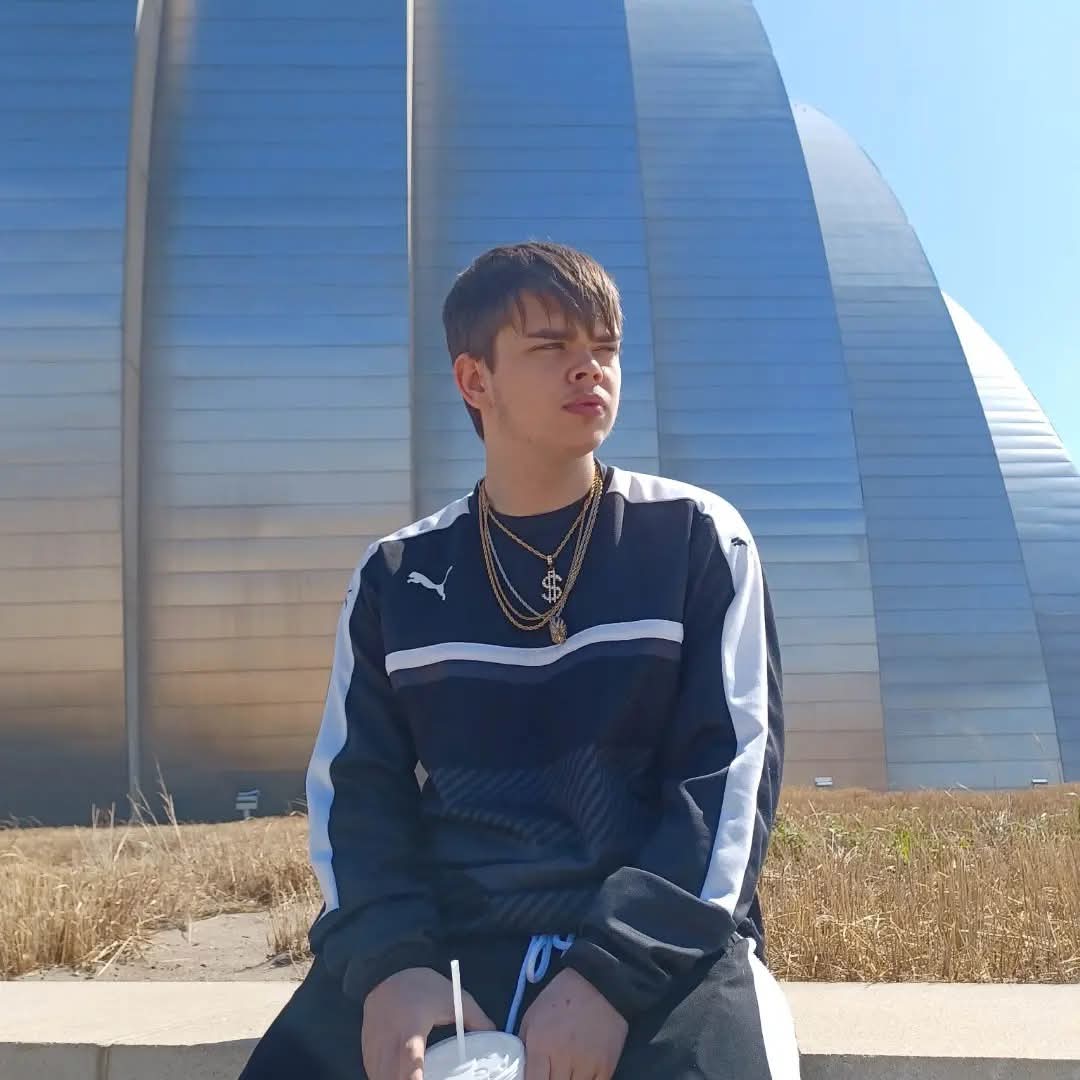 Young man sitting outdoors in front of a modern glass building, wearing a sports jacket and layered necklaces, with sunlight on his face.