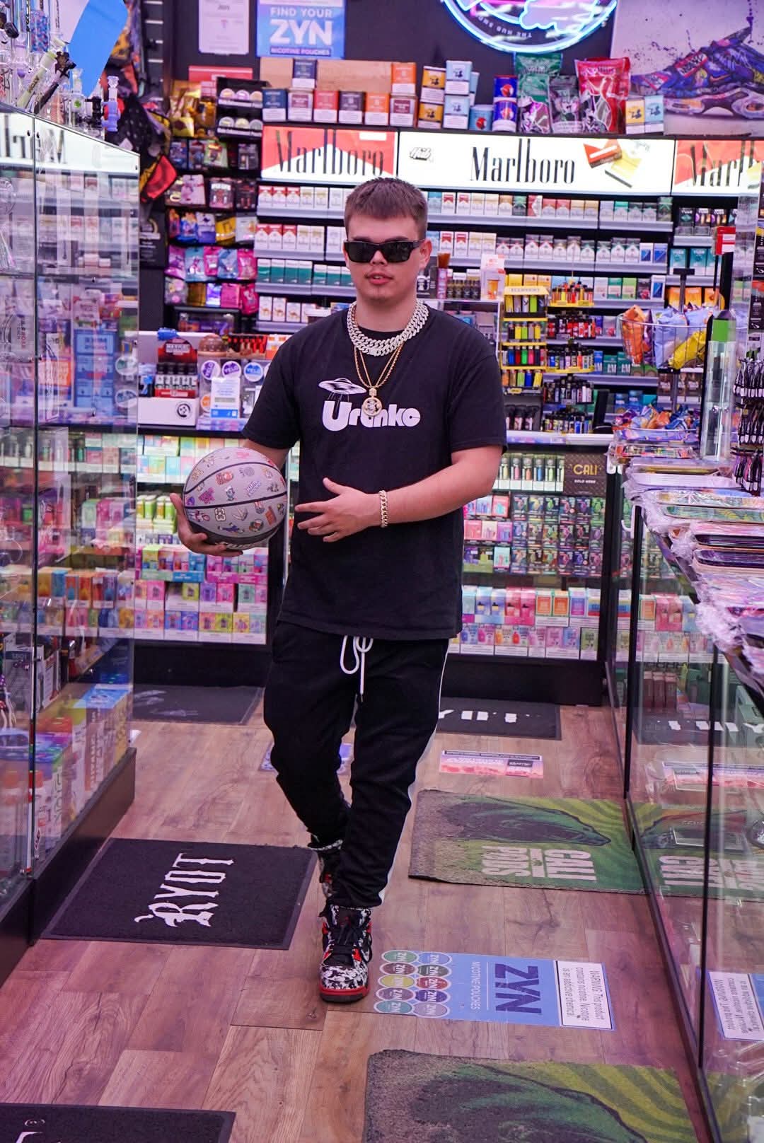 Young person in a store holding a basketball, wearing sunglasses, black t-shirt, and black pants, surrounded by shelves of products.