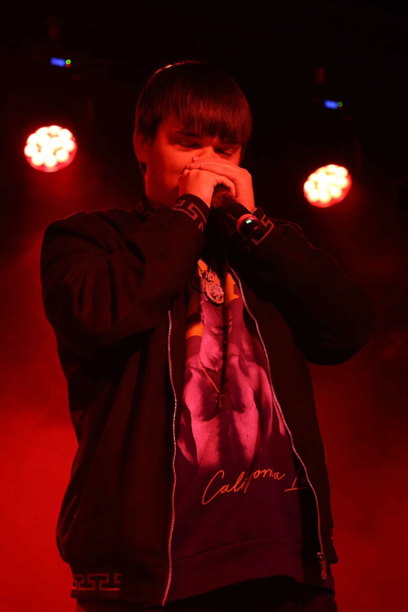 Person with dark hair singing into a microphone on stage with red lighting, wearing a black jacket and graphic T-shirt.