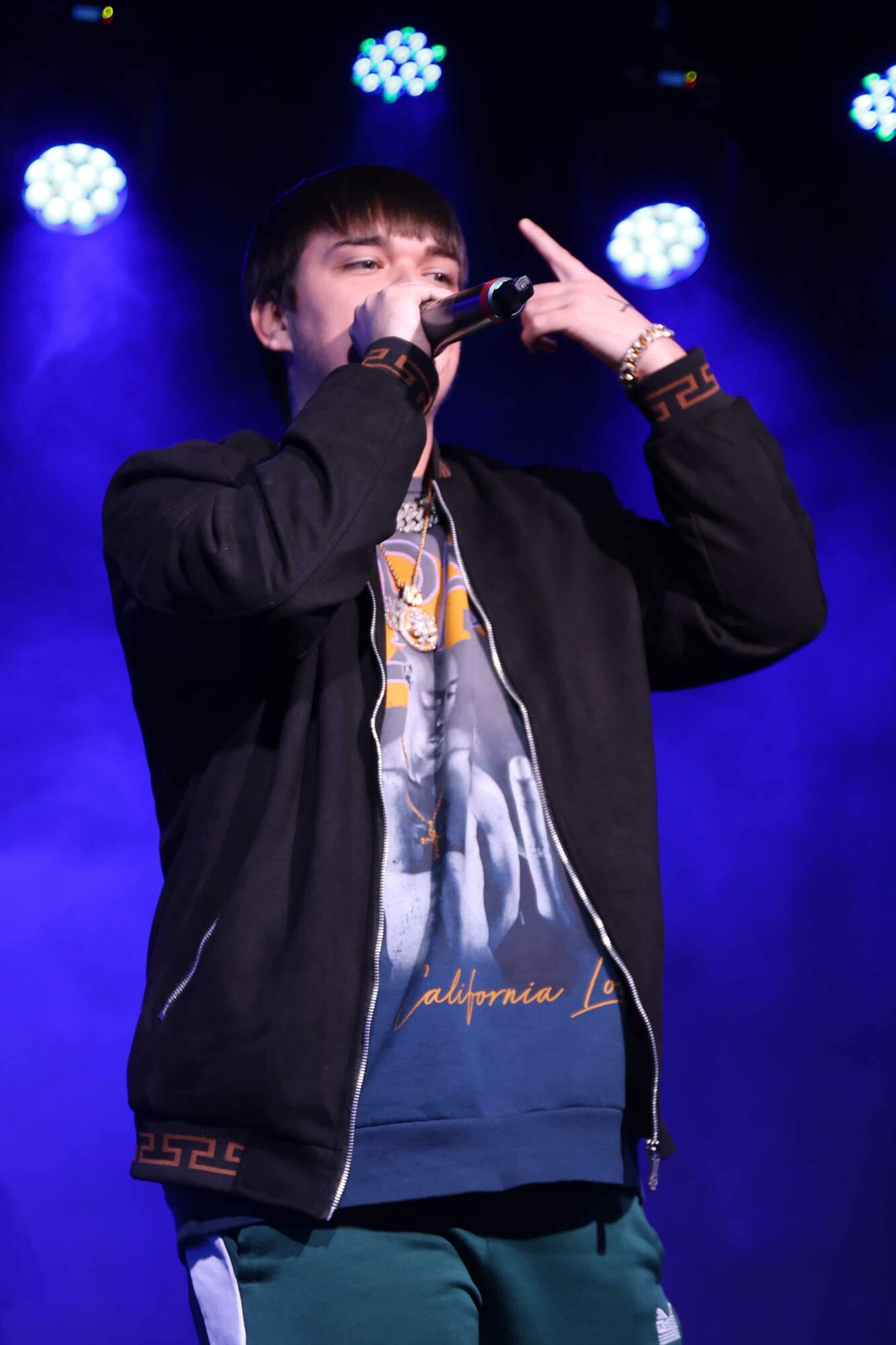 Performer singing into microphone on stage with blue lighting, wearing a black jacket and graphic t-shirt.