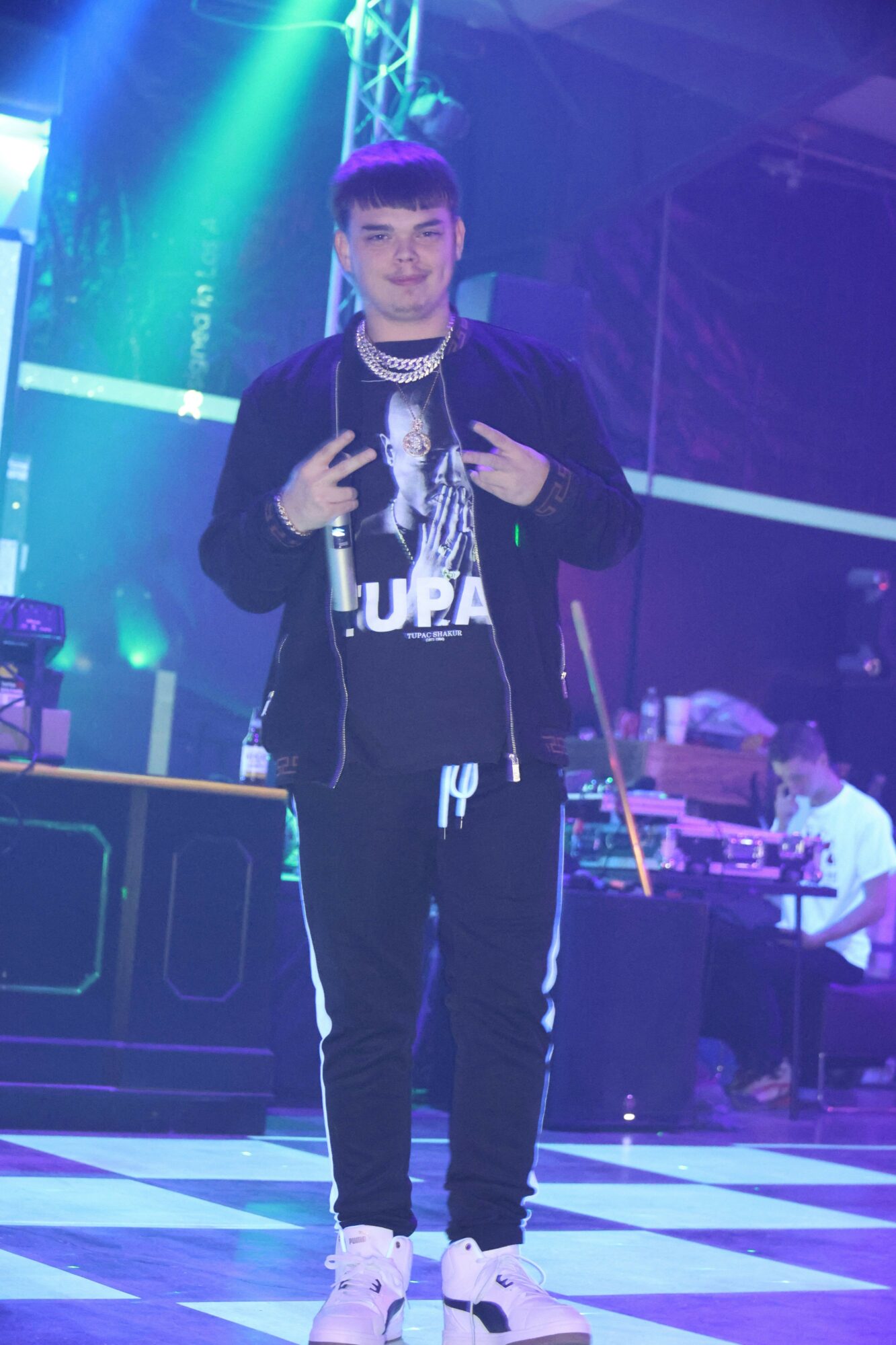 Young man with purple hair holding a microphone on stage with colorful lights and a checkered floor.