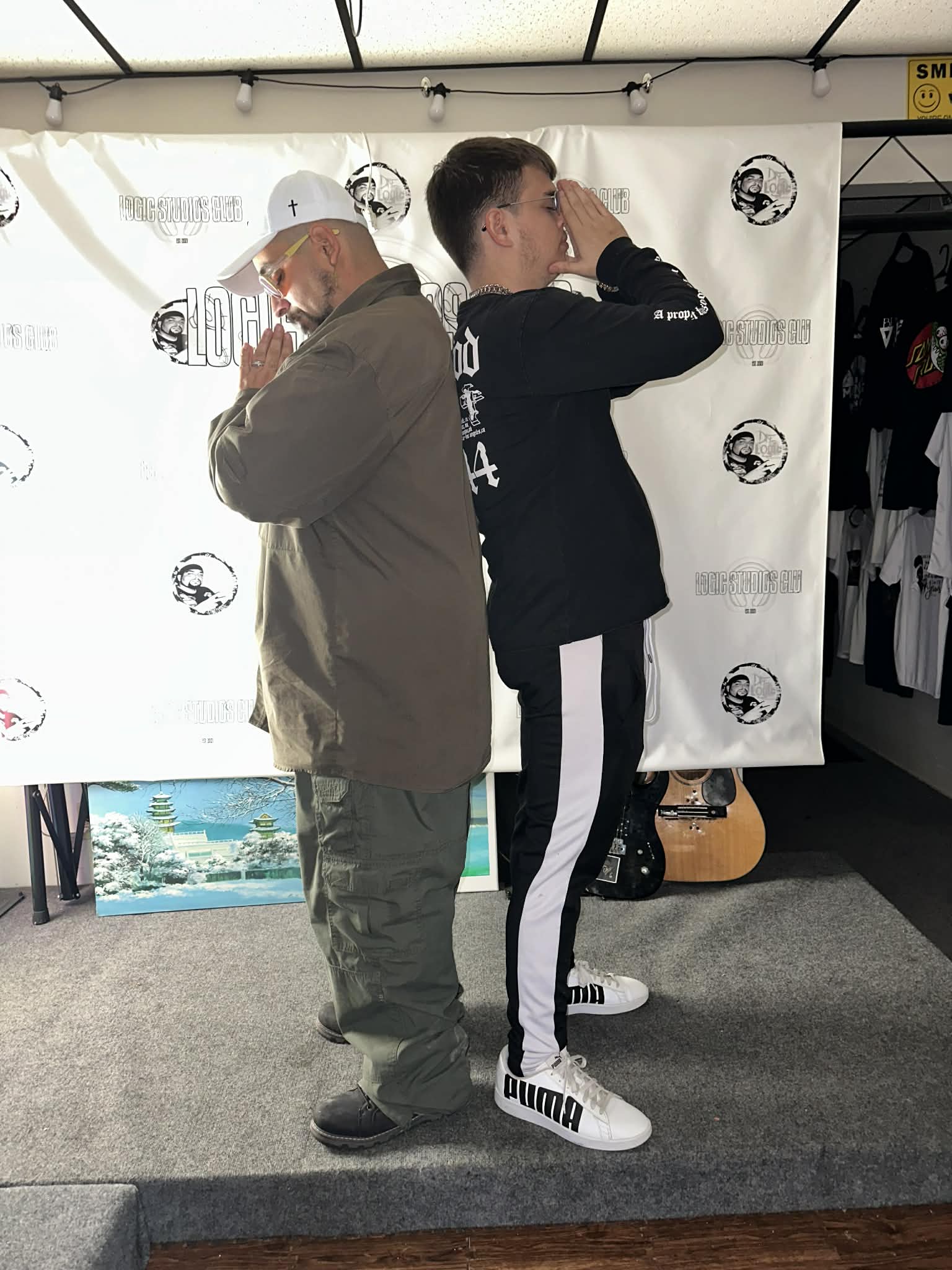 Two men stand back-to-back with arms crossed, facing opposite directions, in front of a white backdrop with logos.