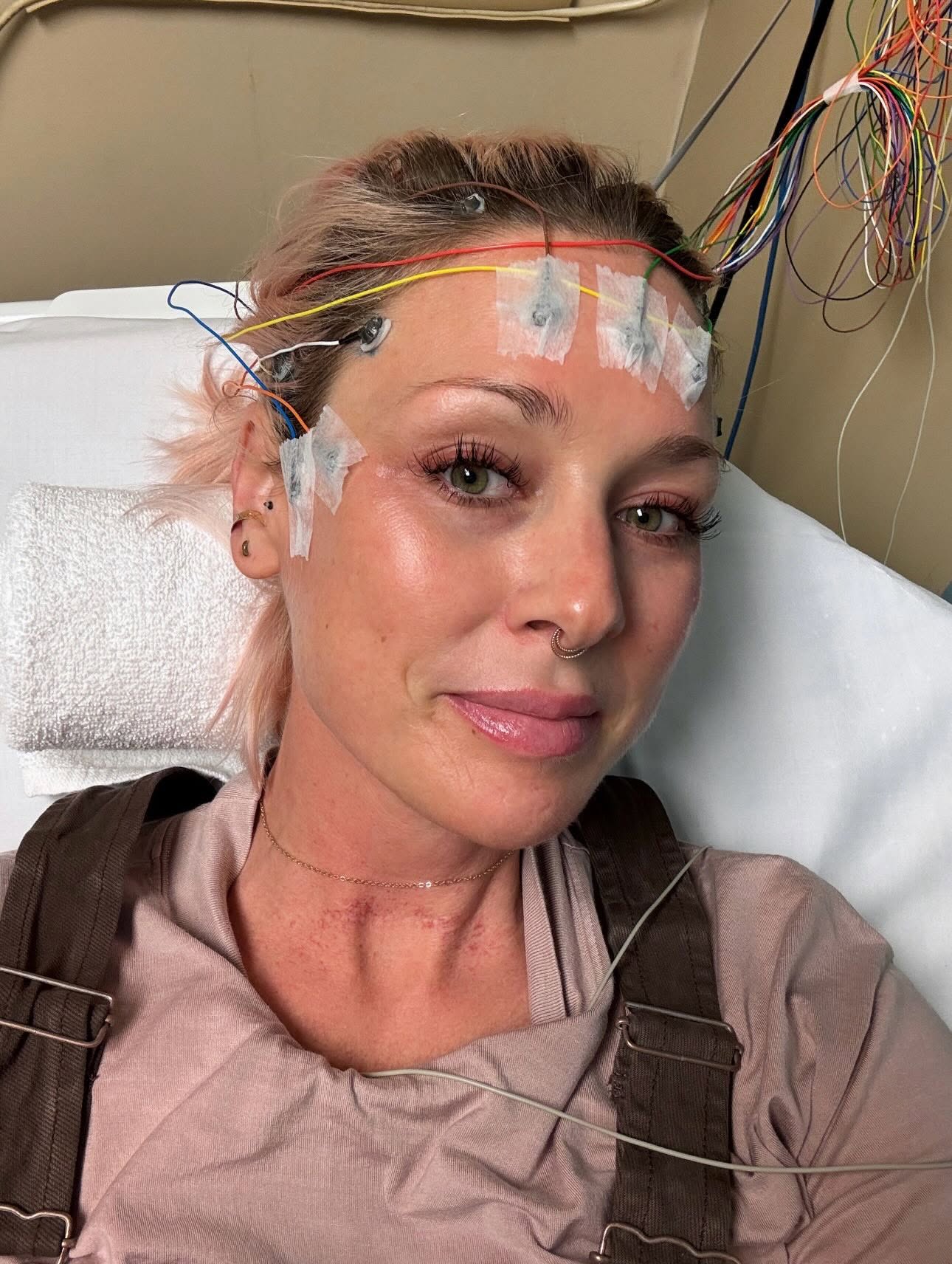 Woman with medical electrodes and wires on her forehead, lying in a hospital bed, smiling gently.