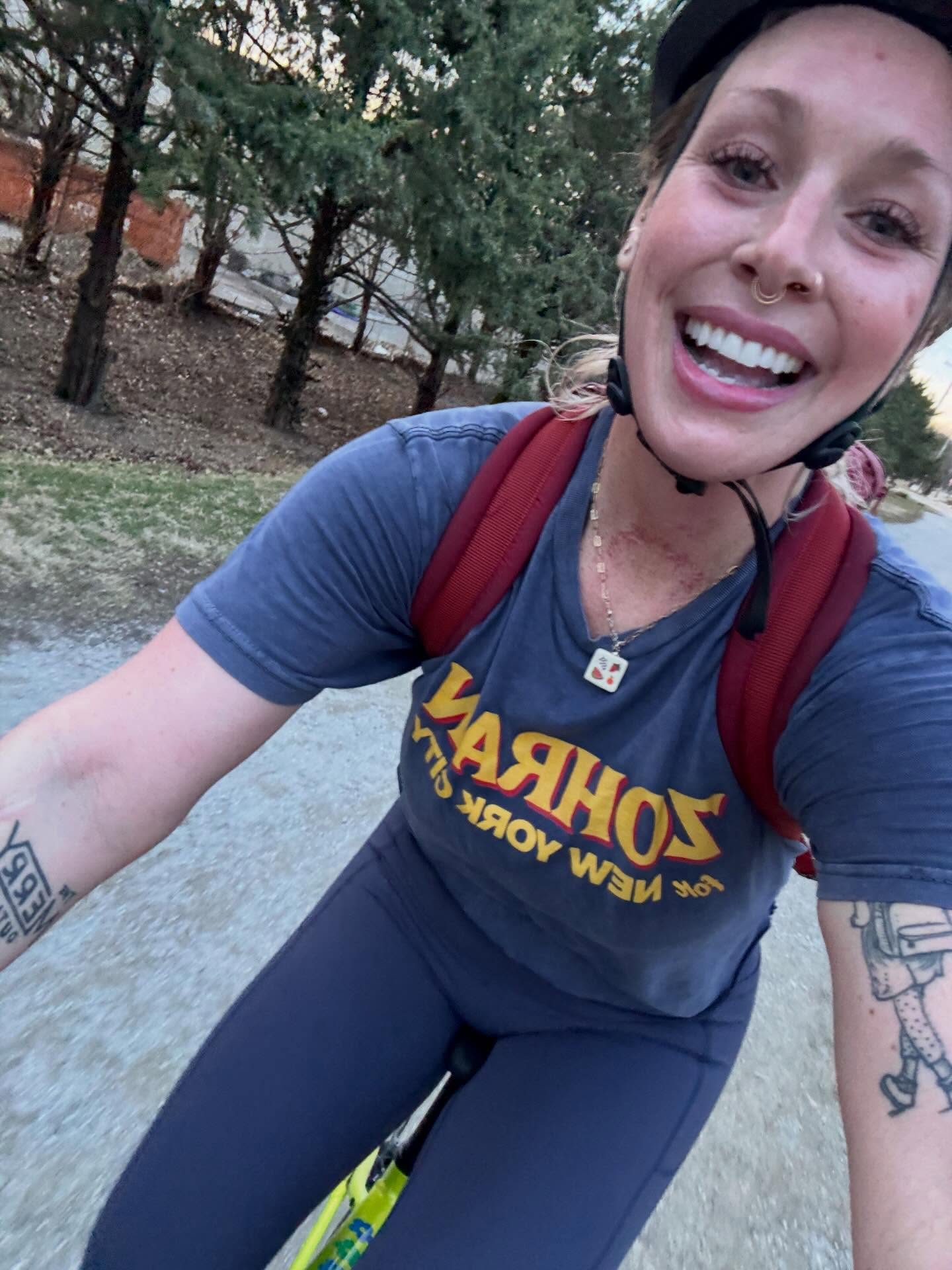 Woman smiling outdoors, wearing a blue shirt with yellow text, a black cap, and a red backpack, with trees in background.