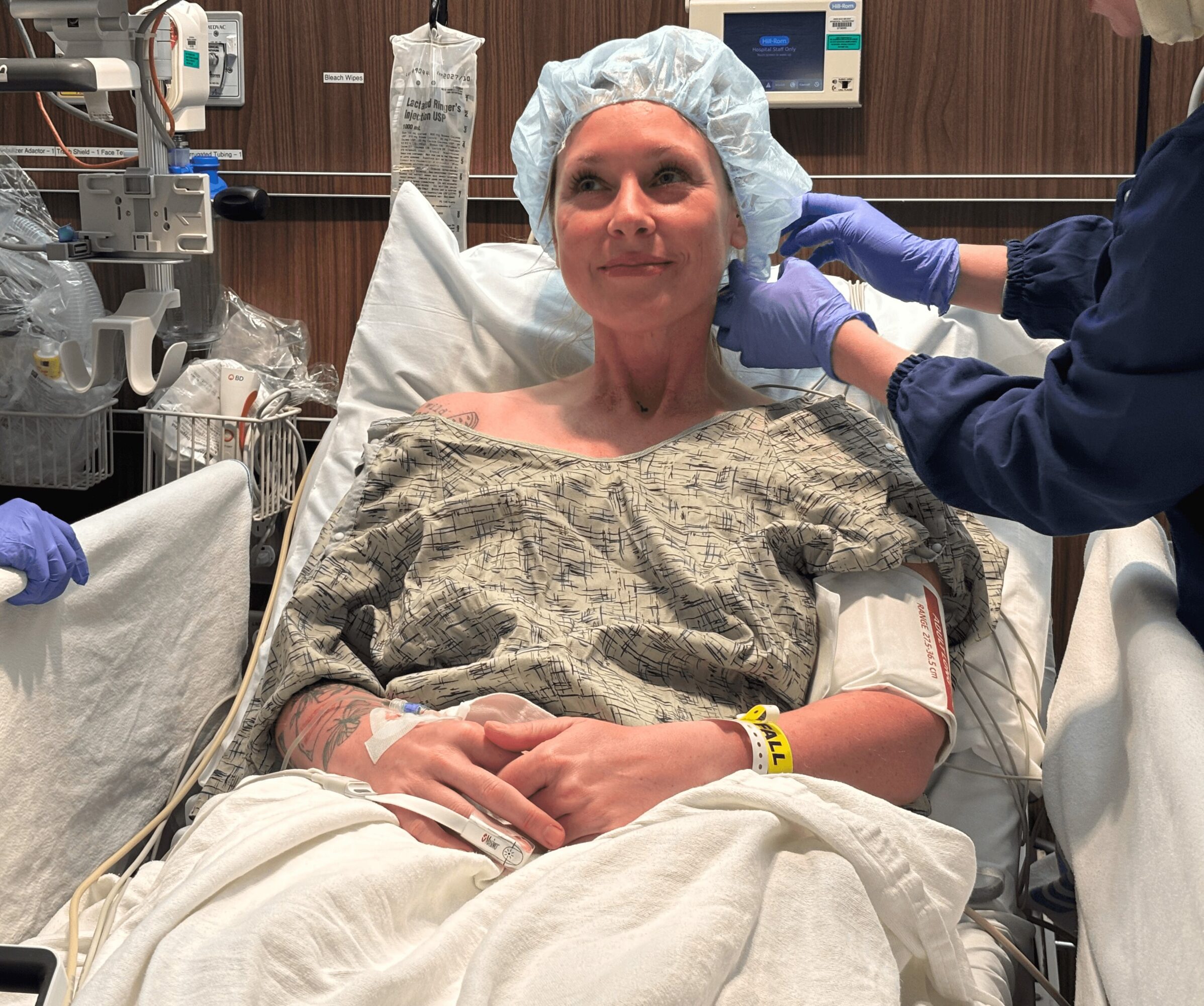 Woman in hospital bed with medical staff preparing her for a procedure, wearing a hair cap and hospital gown.
