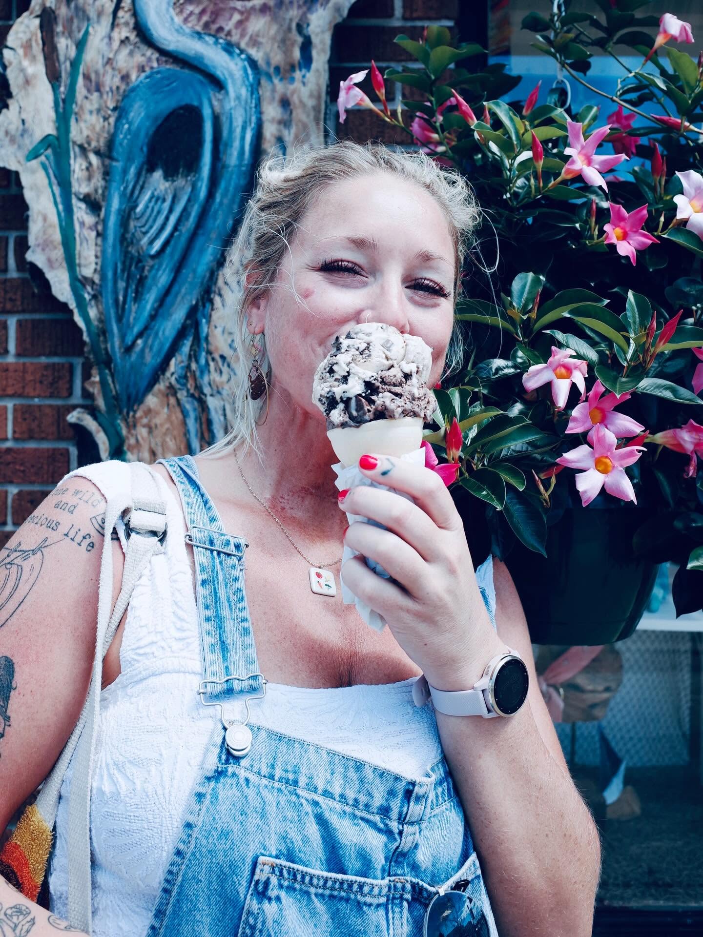 Woman with blonde hair holding an ice cream cone, standing outdoors near pink flowers and a blue mural.