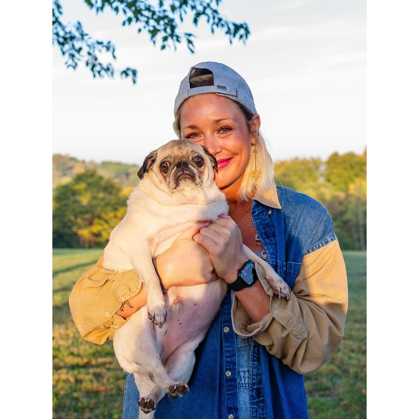 Person holding a pug dog outdoors with trees and sky in background, smiling, wearing a cap and denim jacket.