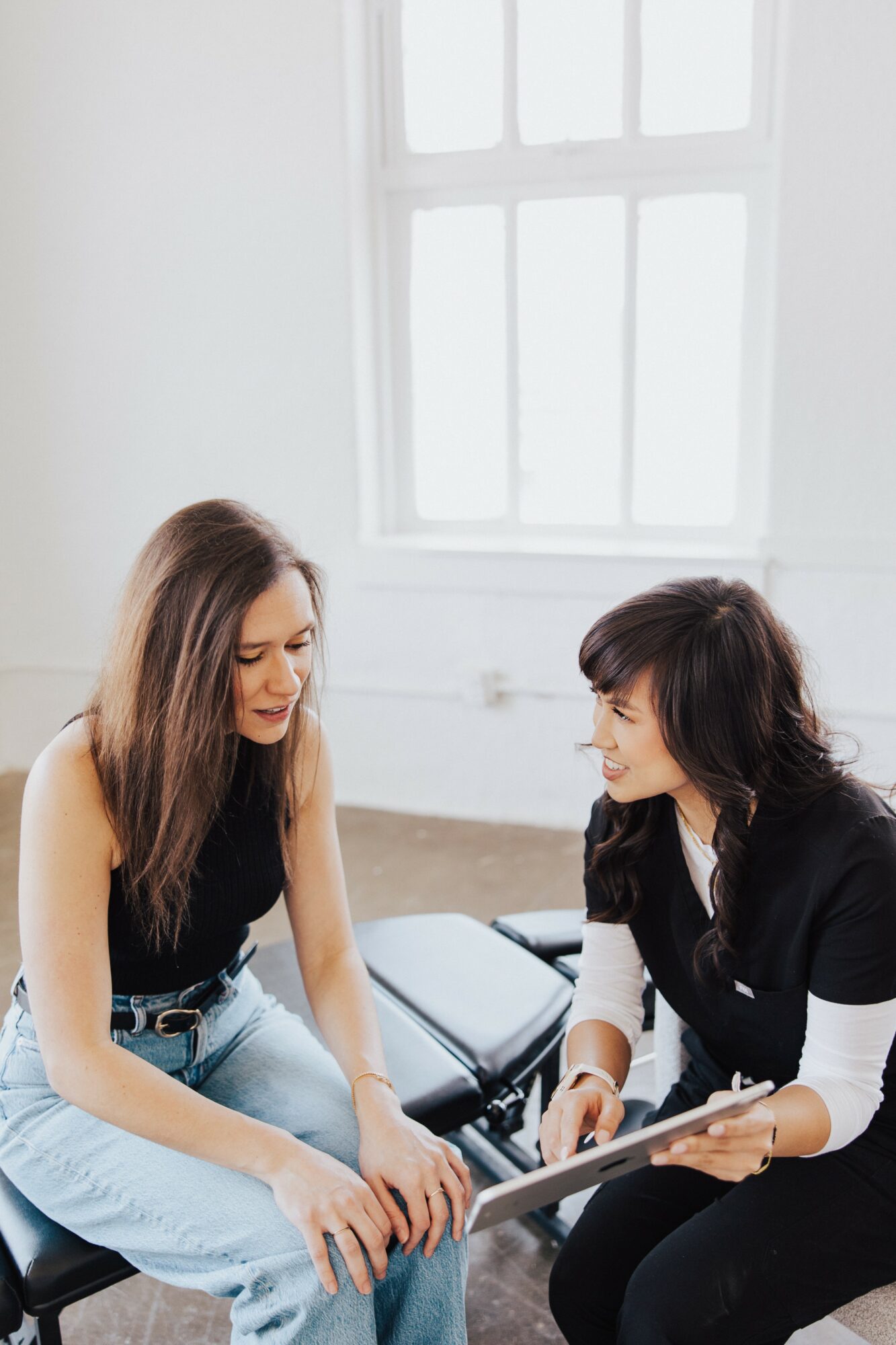 Two women sitting and talking indoors near a window, one holding a tablet, smiling at each other.