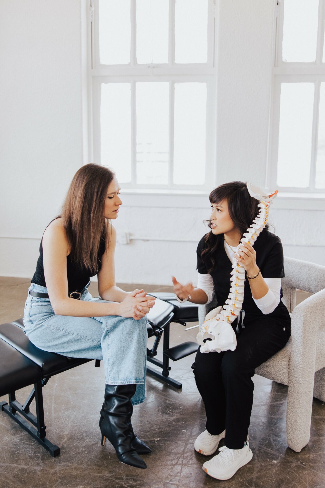 Two women sit and talk in a bright room with large windows, one holding a model of a human spine.